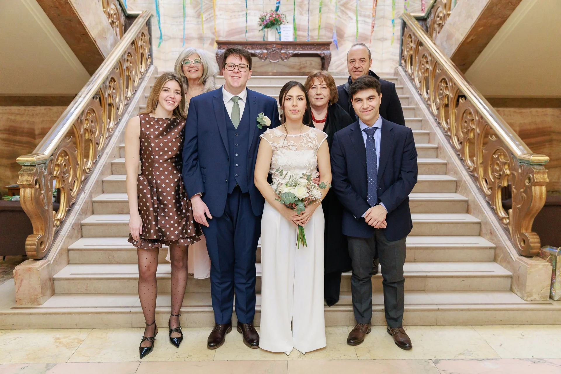 Group of eight people standing on a staircase, dressed in formal attire, with a woman in white holding flowers in the center, celebrating a wedding or special event.