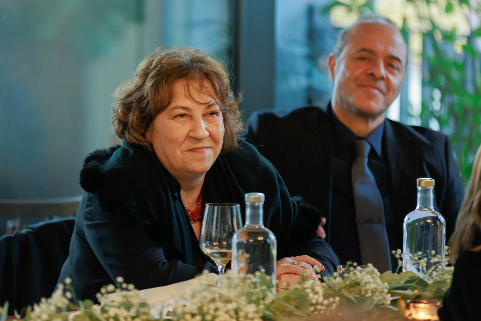 A woman and a man sitting at a table during a formal event. The woman is smiling subtly with her hands clasped in front of her, and the man is smiling broadly in the background. The table has a glass bottle, a wine glass, and floral decorations.