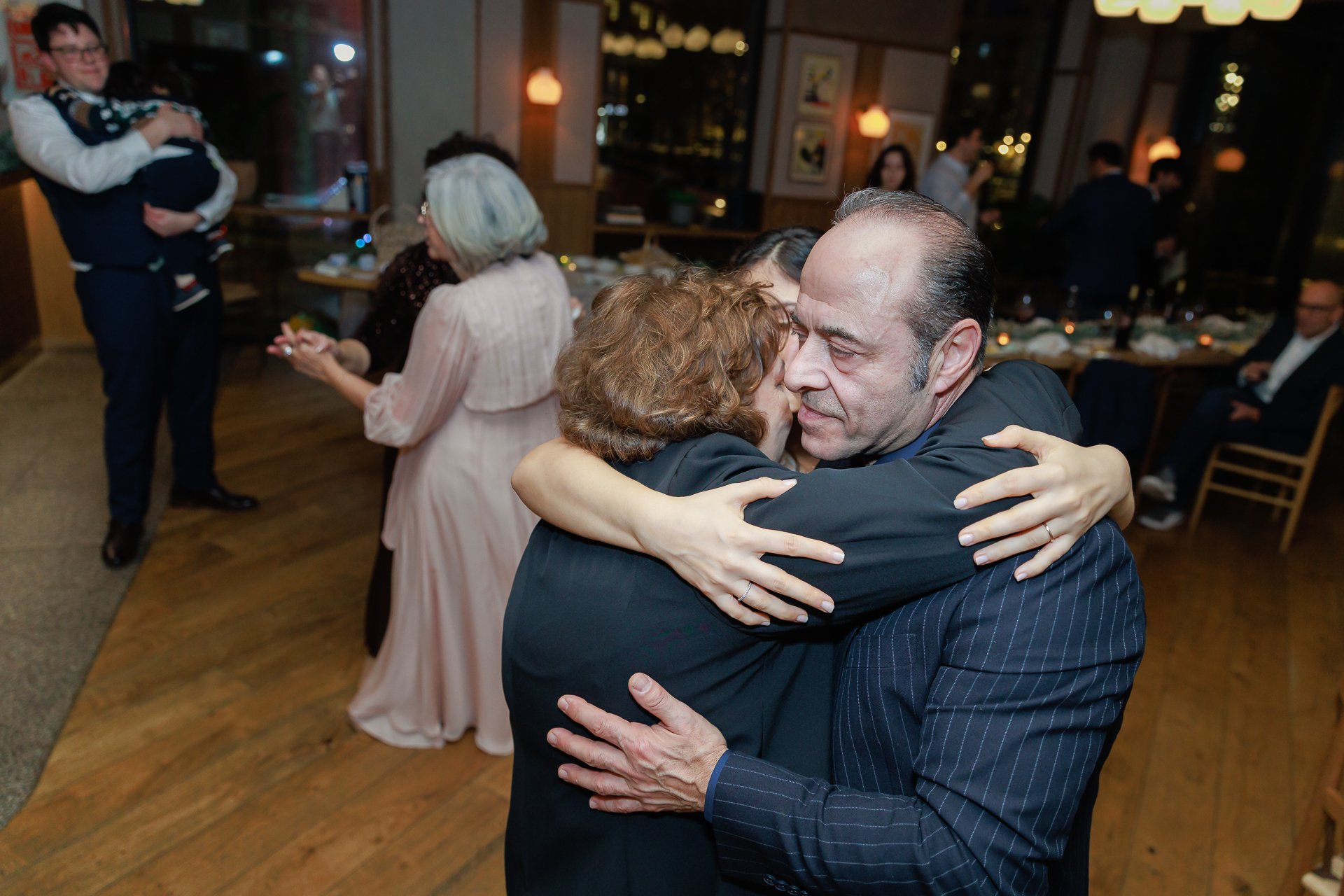 A man and woman share an embrace at a social event, with other guests in the background.