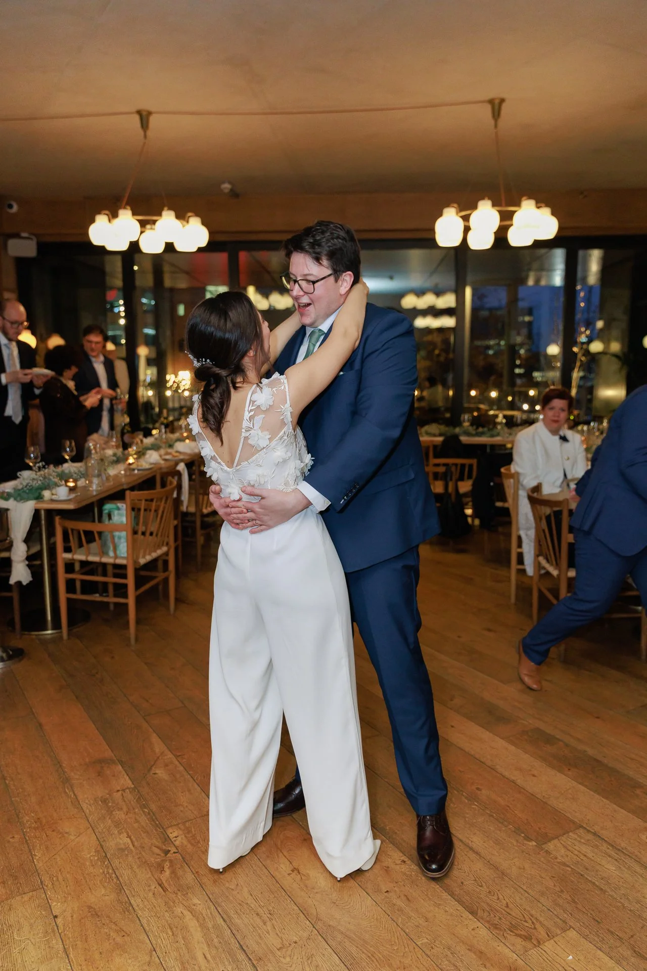 A couple dancing at a wedding reception, the woman in a white dress with flower details and the man in a blue suit, smiling and looking happy.