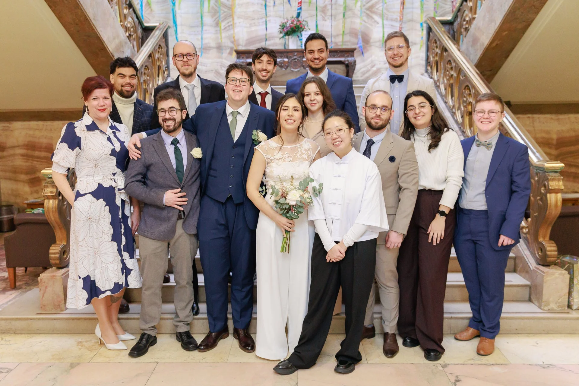 Group of people at a wedding celebration, posing on a staircase with colorful streamers and flowers in the background.
