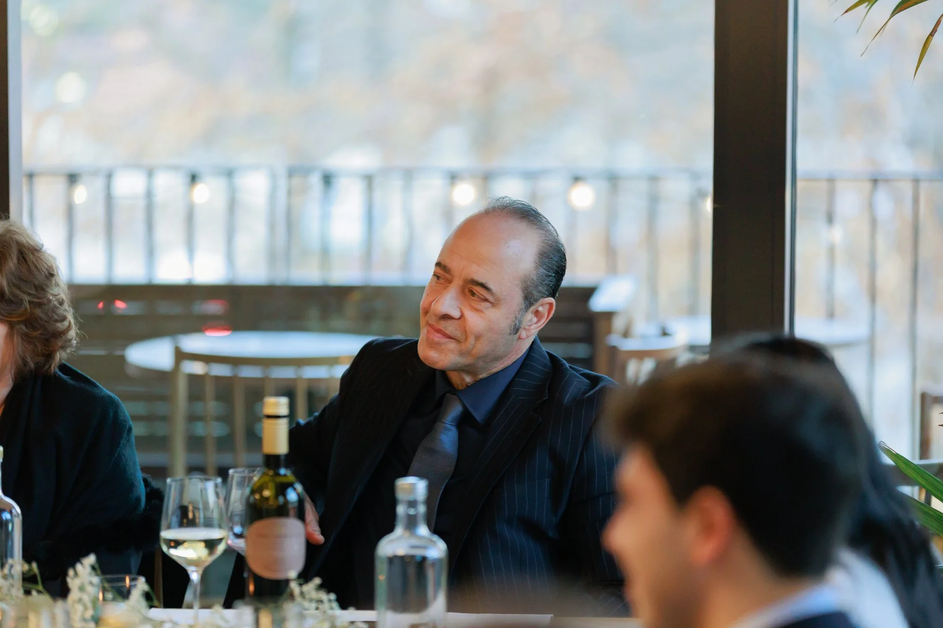 Man in a suit and tie sitting at a table with wine and water bottles, in a room with large windows and natural light.