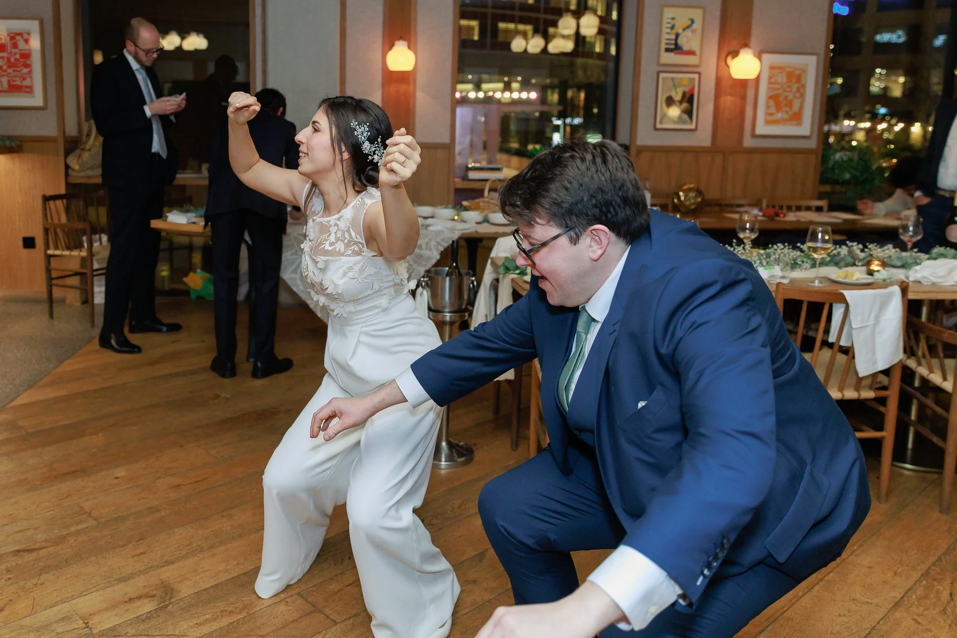 A woman in a white dress and a man in a blue suit dancing and having fun at a celebration or wedding reception, with other guests and decorated tables in the background.