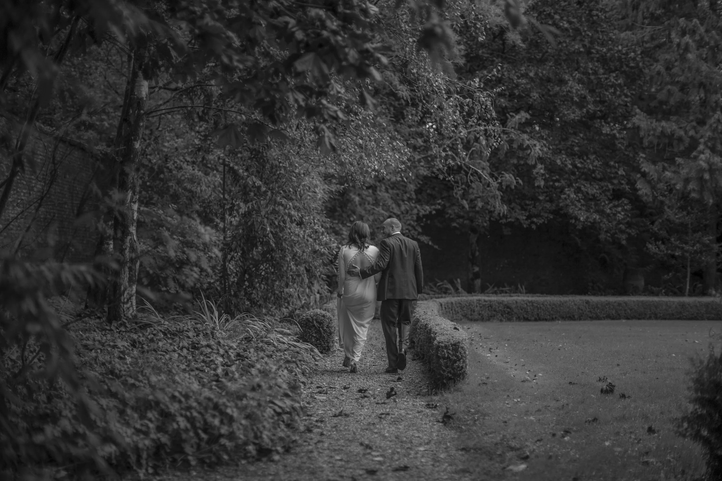 A black and white photo of a couple walking hand in hand along a garden path surrounded by trees and bushes.