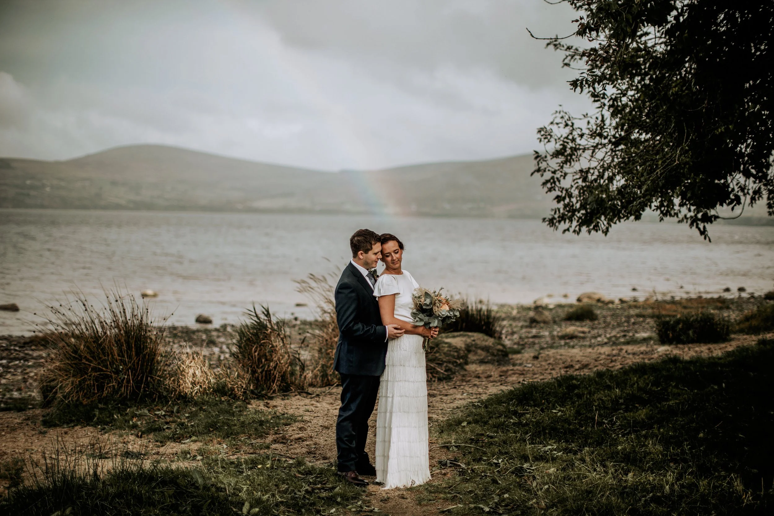 A bride and groom stand close together by a lakeshore, with the bride holding a bouquet of flowers. They are dressed in wedding attire, with a cloudy sky and a faint rainbow in the background.