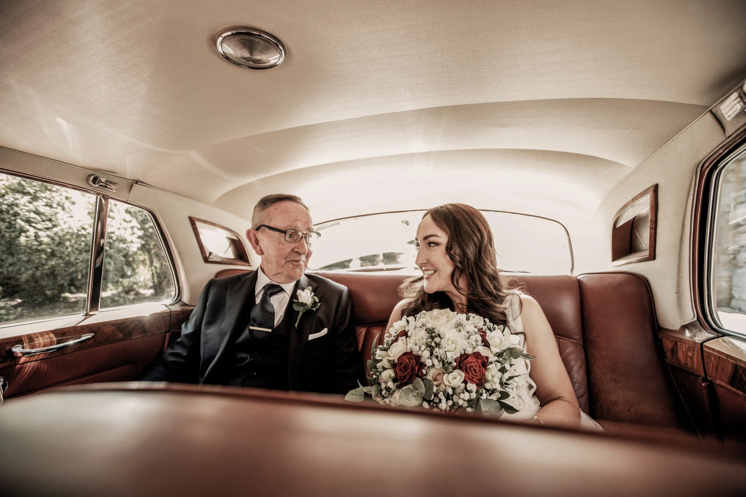 A bride smiling and holding a bouquet of roses and white flowers, sitting inside a vintage car with an older man in a suit, likely her father, looking at her.
