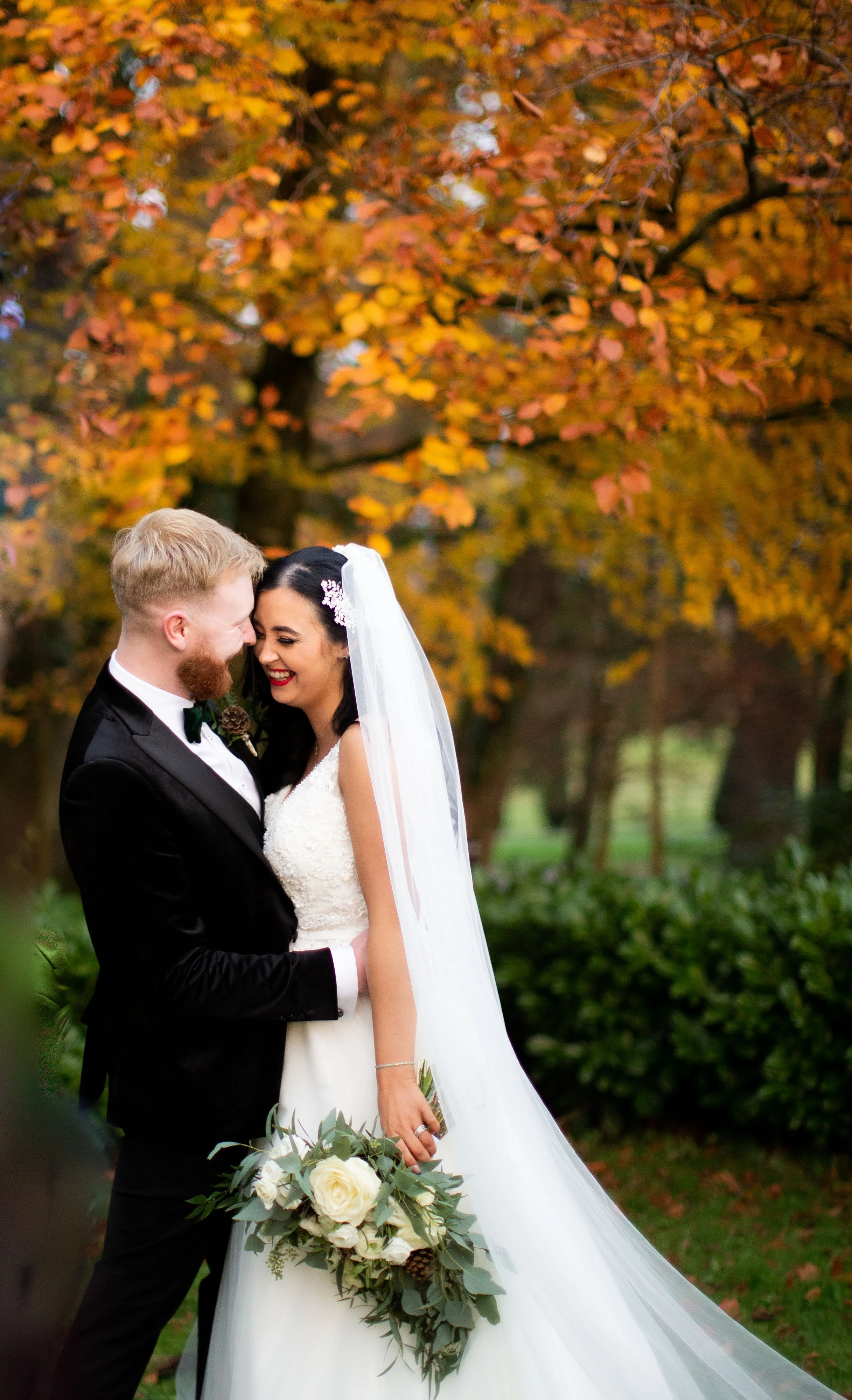 A newlywed couple, a man in a black tuxedo and woman in a white wedding gown with a veil, sharing a joyful moment outdoors with autumn foliage in the background.