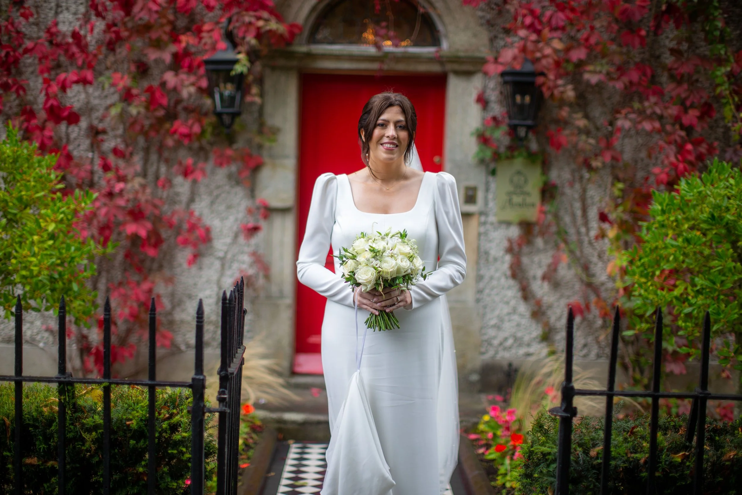 A woman in a white dress holding a bouquet of white roses stands in front of a red door covered with red and green vines, with black lanterns on either side of the door, and a garden with pink and red flowers surrounding her.