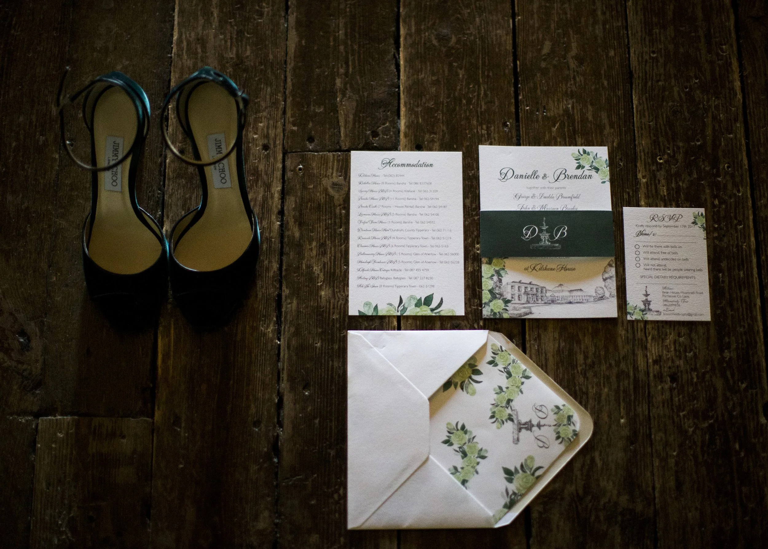 Black high heels, wedding invitation, RSVP card, accommodation list, and tray with floral design on dark wooden floor.