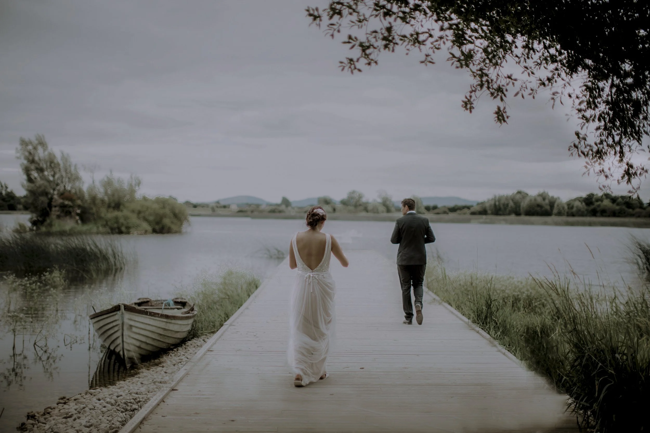 A bride in a white gown and a groom in a dark suit walk along a wooden dock by a lake on a cloudy day.