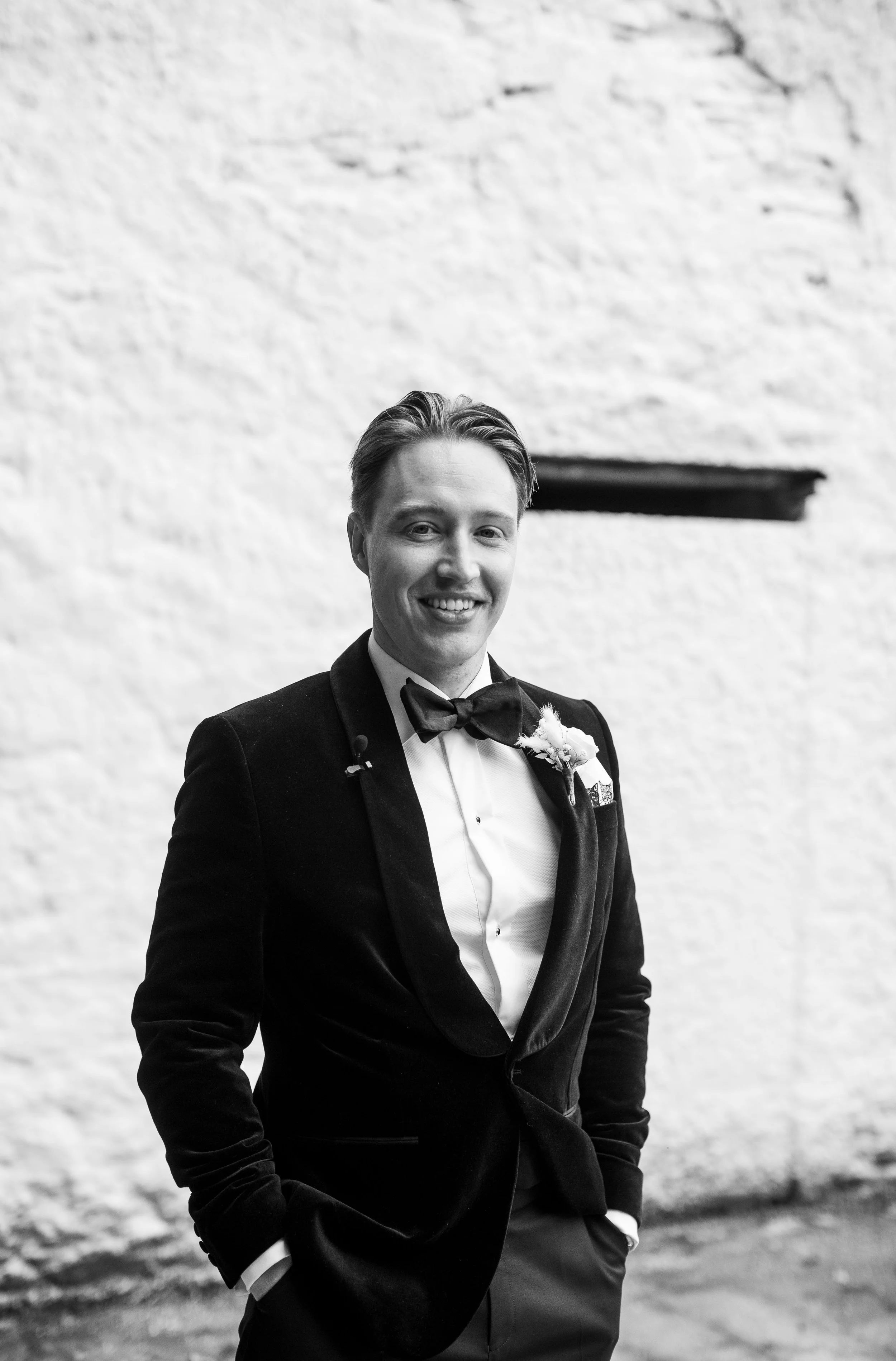 Black and white photo of a smiling young man in formal tuxedo with a bow tie and boutonniere, standing with hands in pockets in front of a textured white wall.