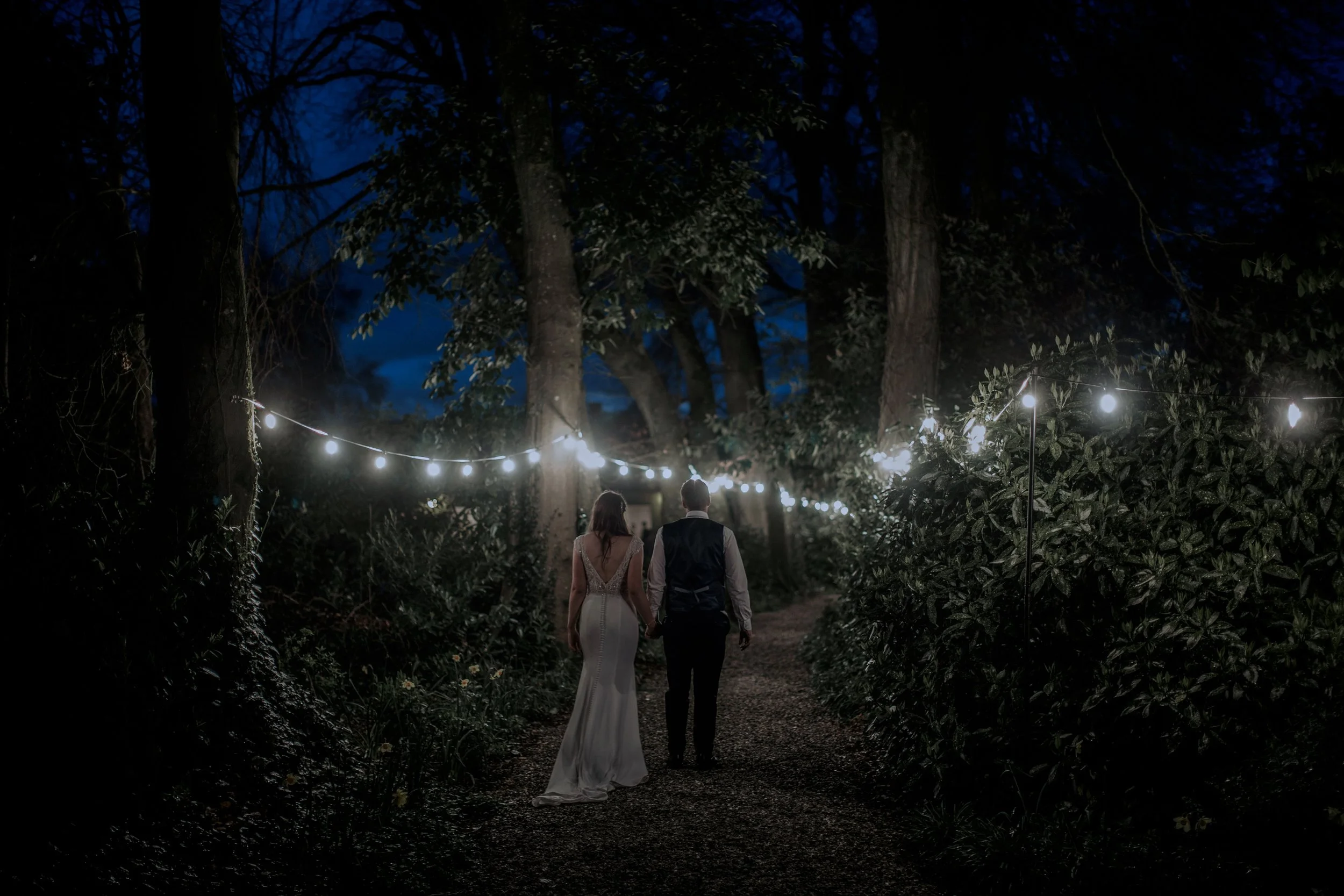 A couple walking hand-in-hand through a wooded area decorated with hanging string lights at night, dressed in wedding attire.