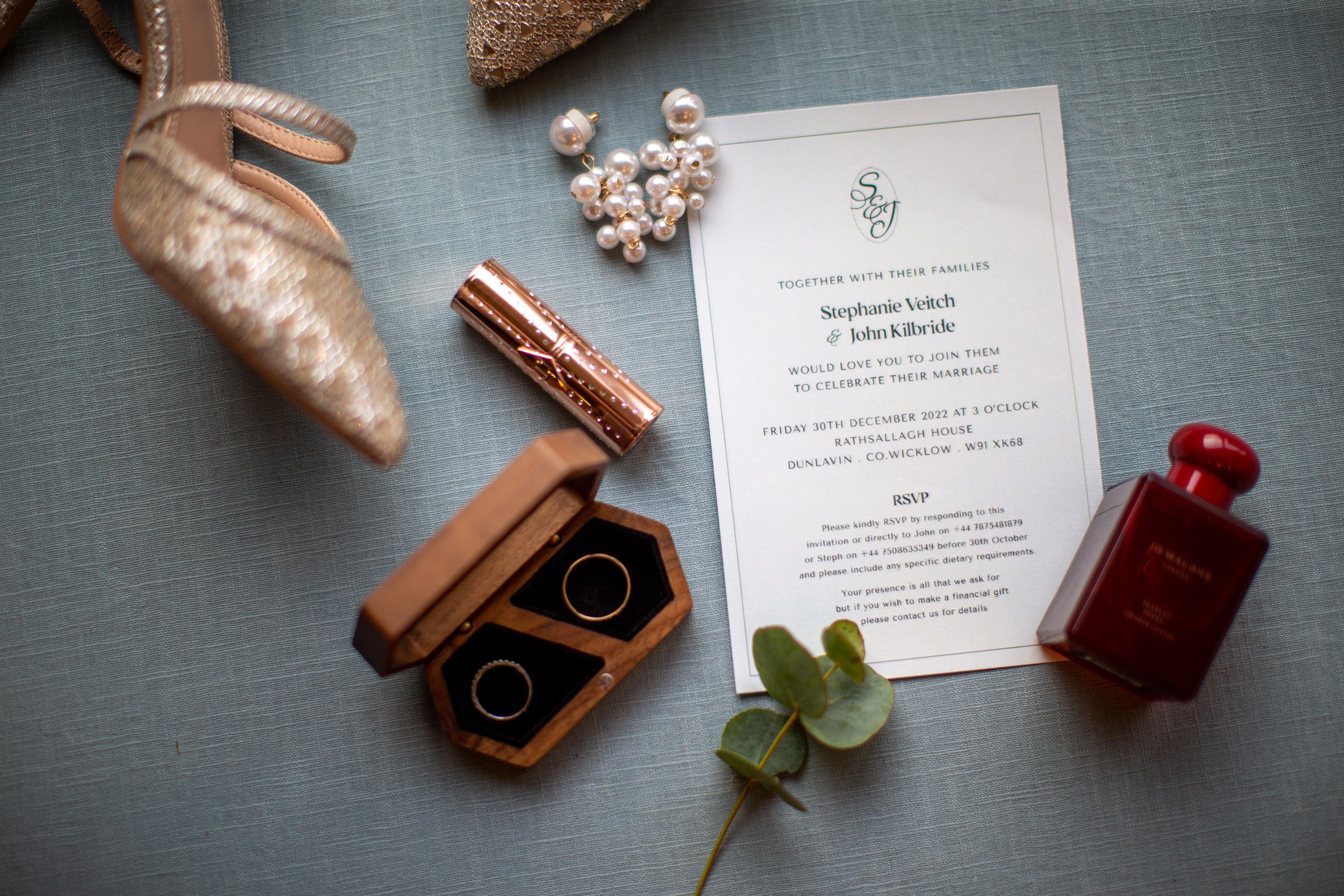 Wedding invitation surrounded by jewelry and accessories including pearl earrings, gold shoes, rings in a wooden box, red perfume, a small wrapped gift, and greenery on a light blue fabric background.