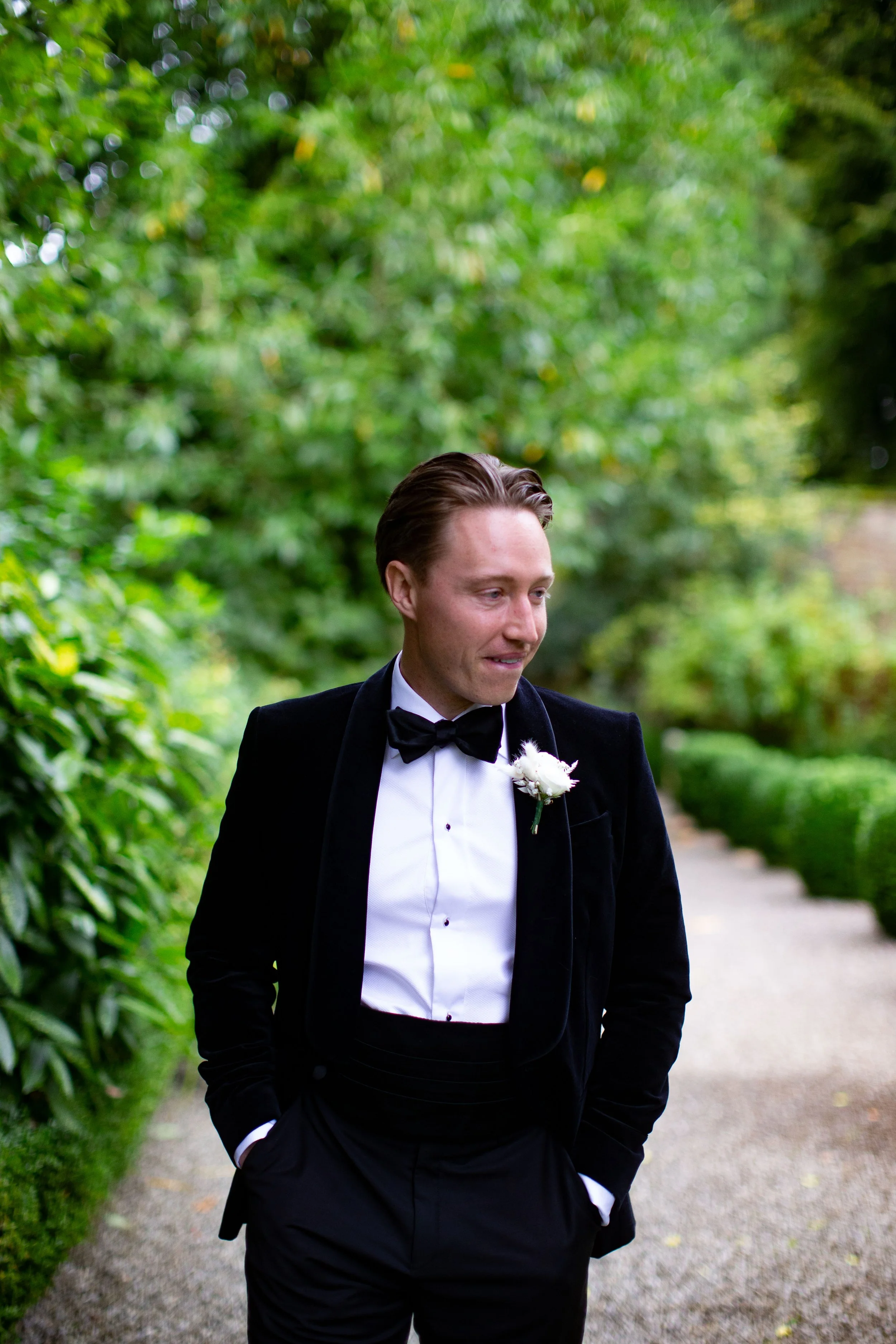 Man in black tuxedo with bow tie and white shirt walking on a gravel path surrounded by lush green trees