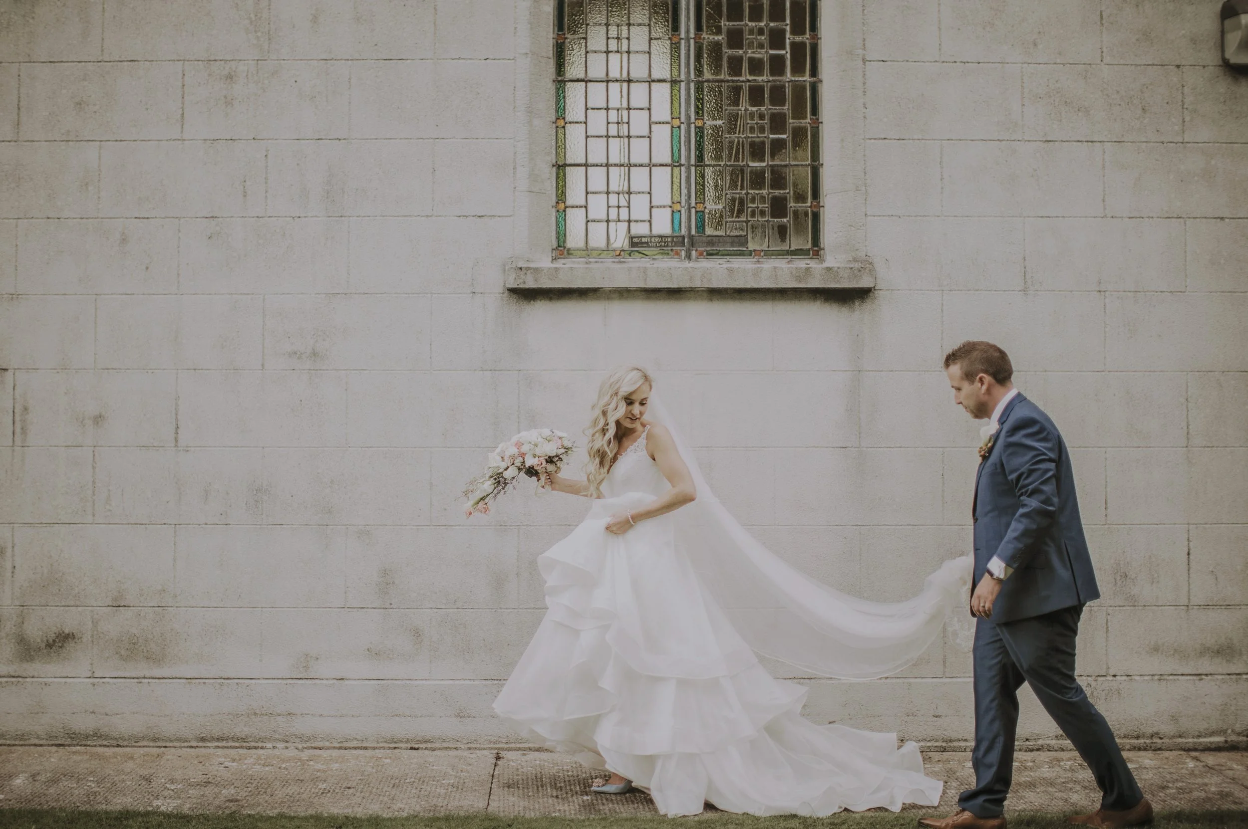 A couple in wedding attire walking along a concrete sidewalk in front of a gray stone wall with a stained glass window. The bride is in a white wedding gown holding a bouquet of flowers, and the groom is in a blue suit.