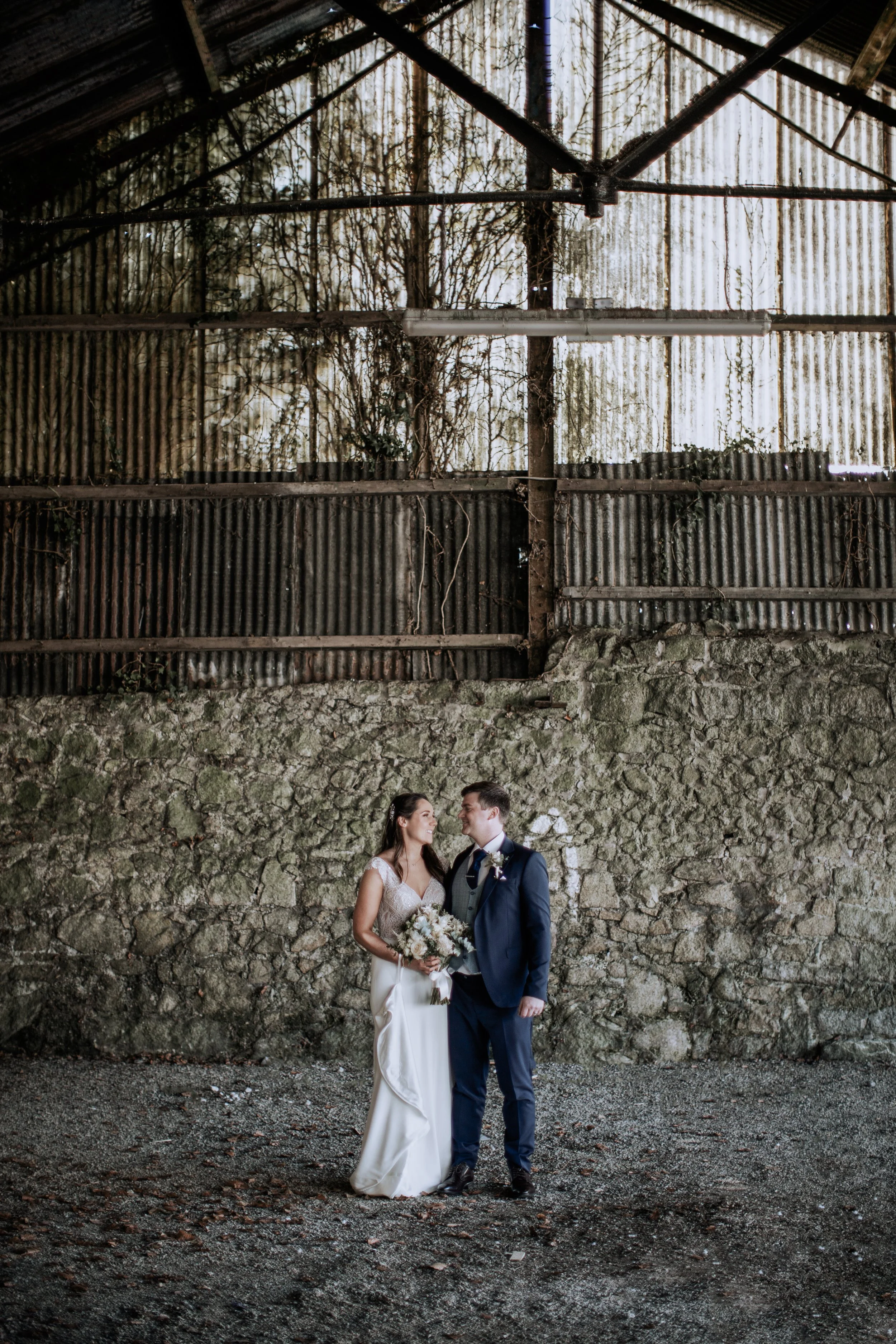 Bride and groom standing together in an industrial, rustic setting with a stone wall and corrugated metal roof, looking at each other, the bride holding a bouquet, and the groom in a navy suit.
