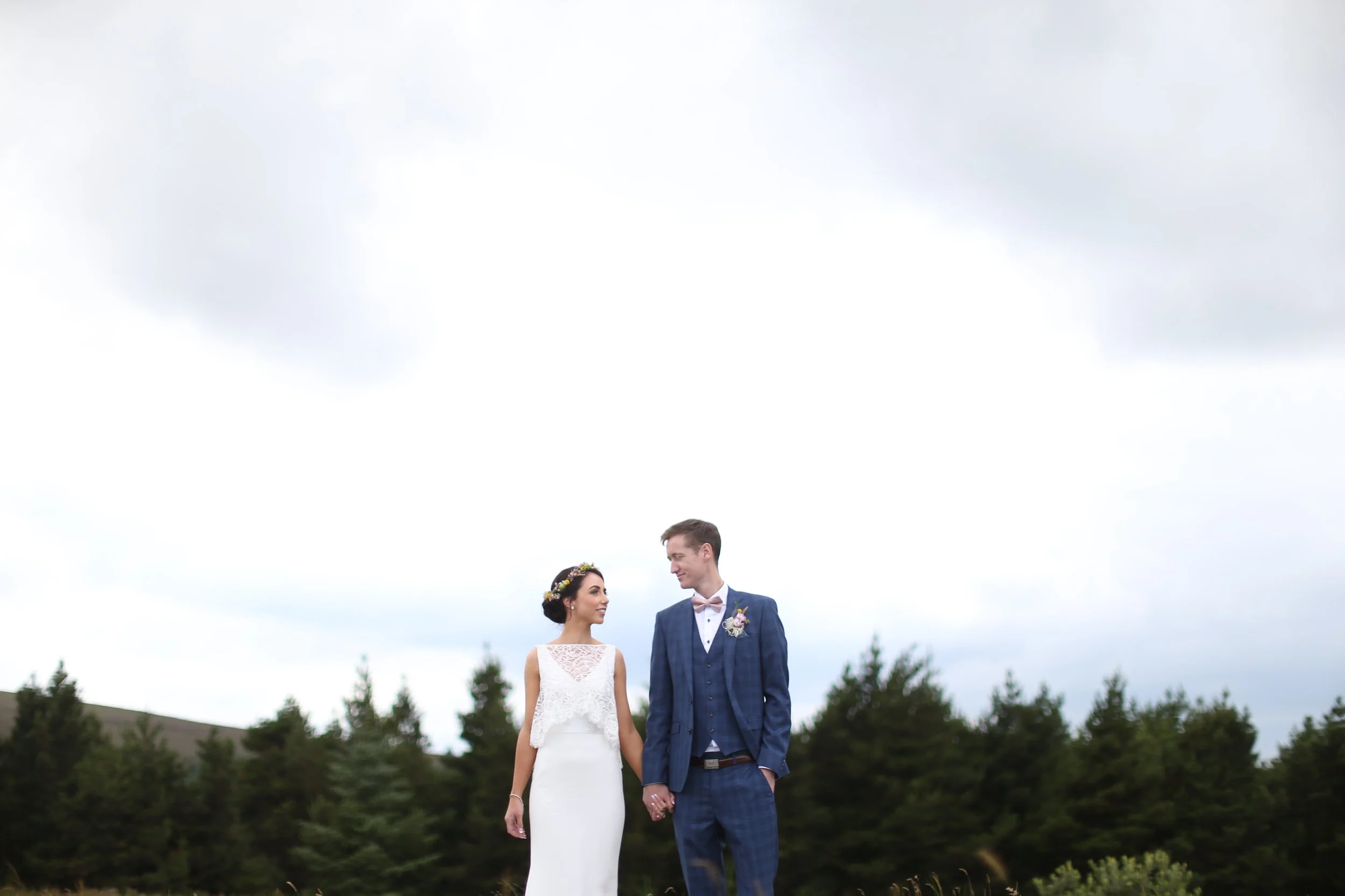 Bride and groom holding hands, standing outdoors in a field with trees, under cloudy sky.