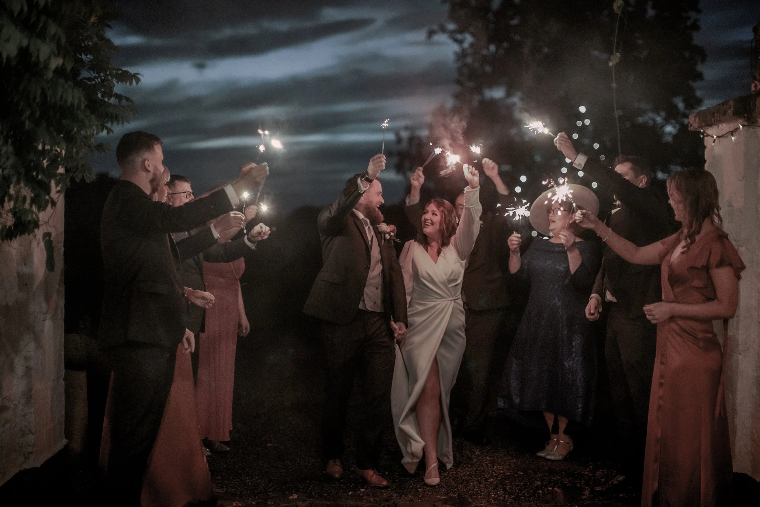 A group of people in formal attire celebrating at night with sparklers, including a woman in a white dress and a man in a suit, standing closely together outdoors under a dark, cloudy sky.