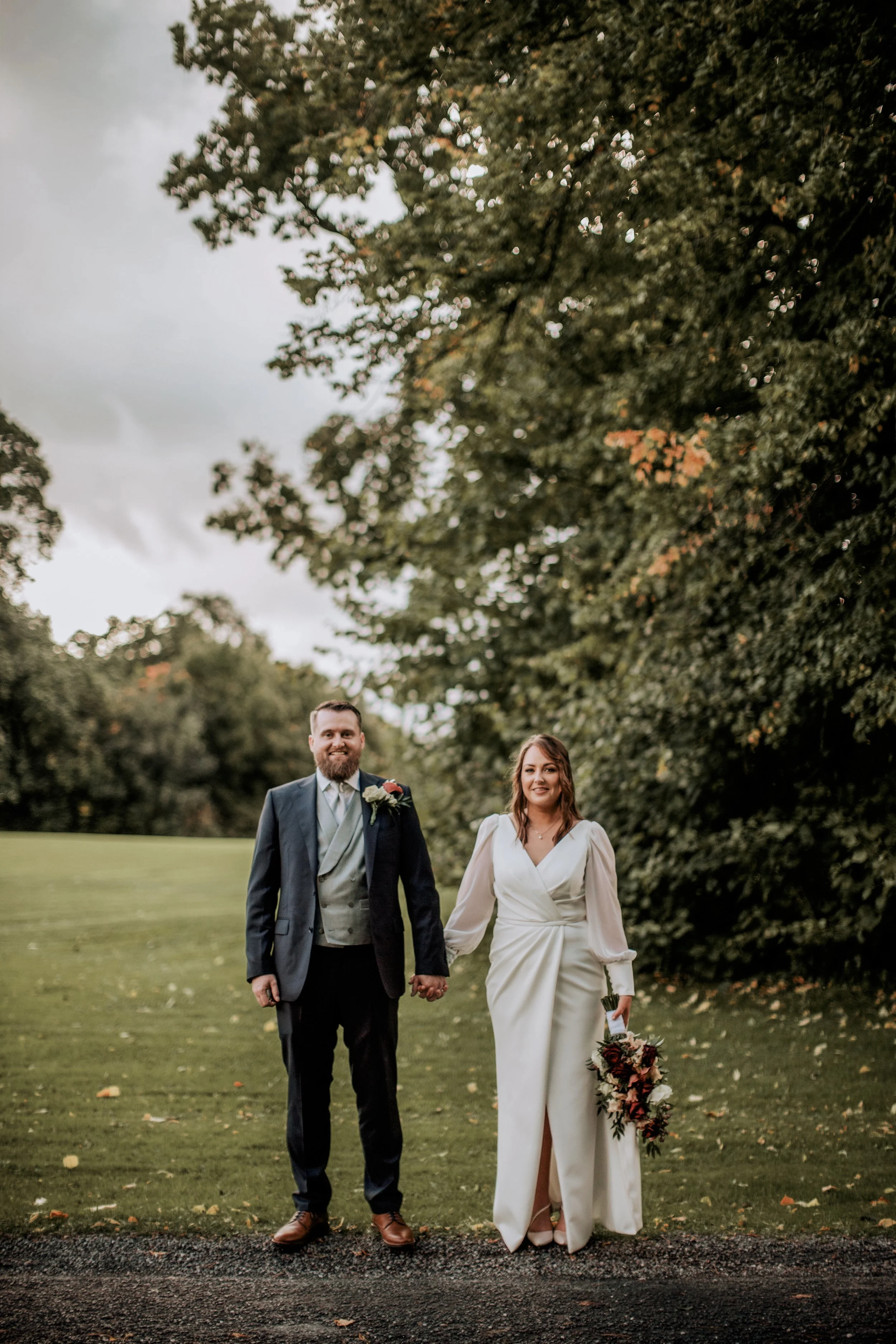 A bride and groom standing outdoors on a grassy area holding hands, with trees in the background. The groom is wearing a dark suit with a boutonniere, and the bride is wearing a white wedding dress and holding a bouquet of flowers.