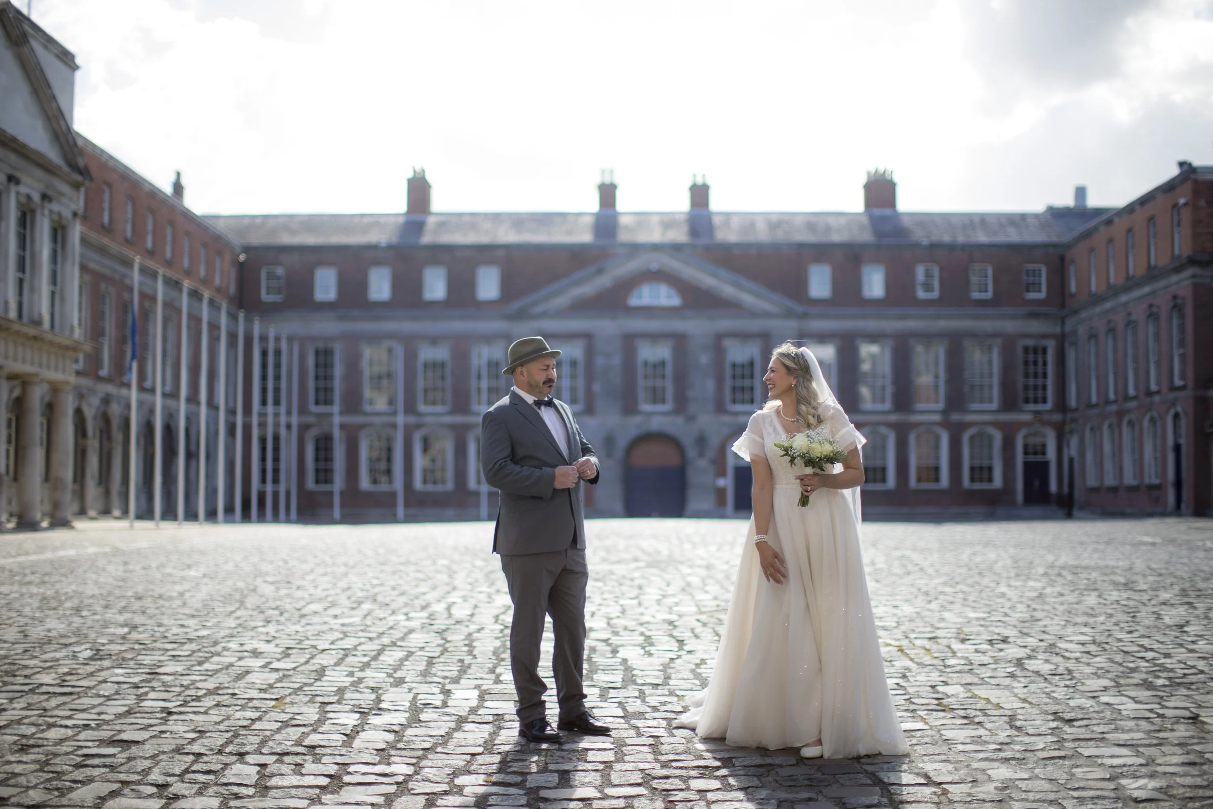 Bride and groom standing outside on a cobblestone courtyard in front of a large historic brick building, smiling at each other. The bride is wearing a white wedding dress holding a bouquet, and the groom is dressed in a gray suit with a bow tie and h