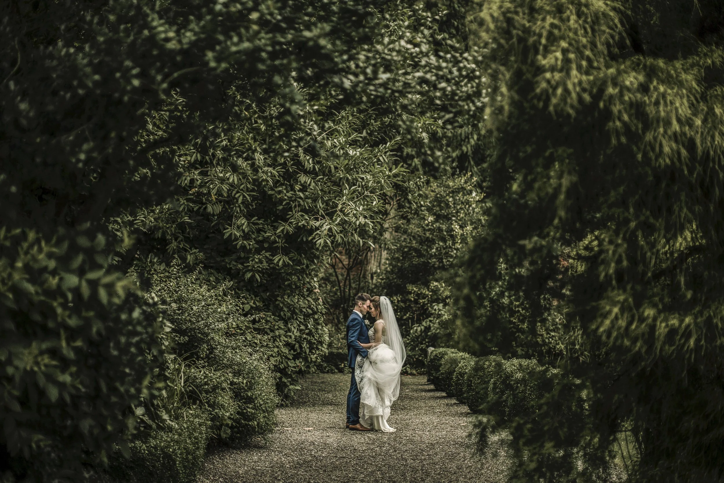 A bride and groom standing close and holding hands in a lush green garden surrounded by dense bushes and trees