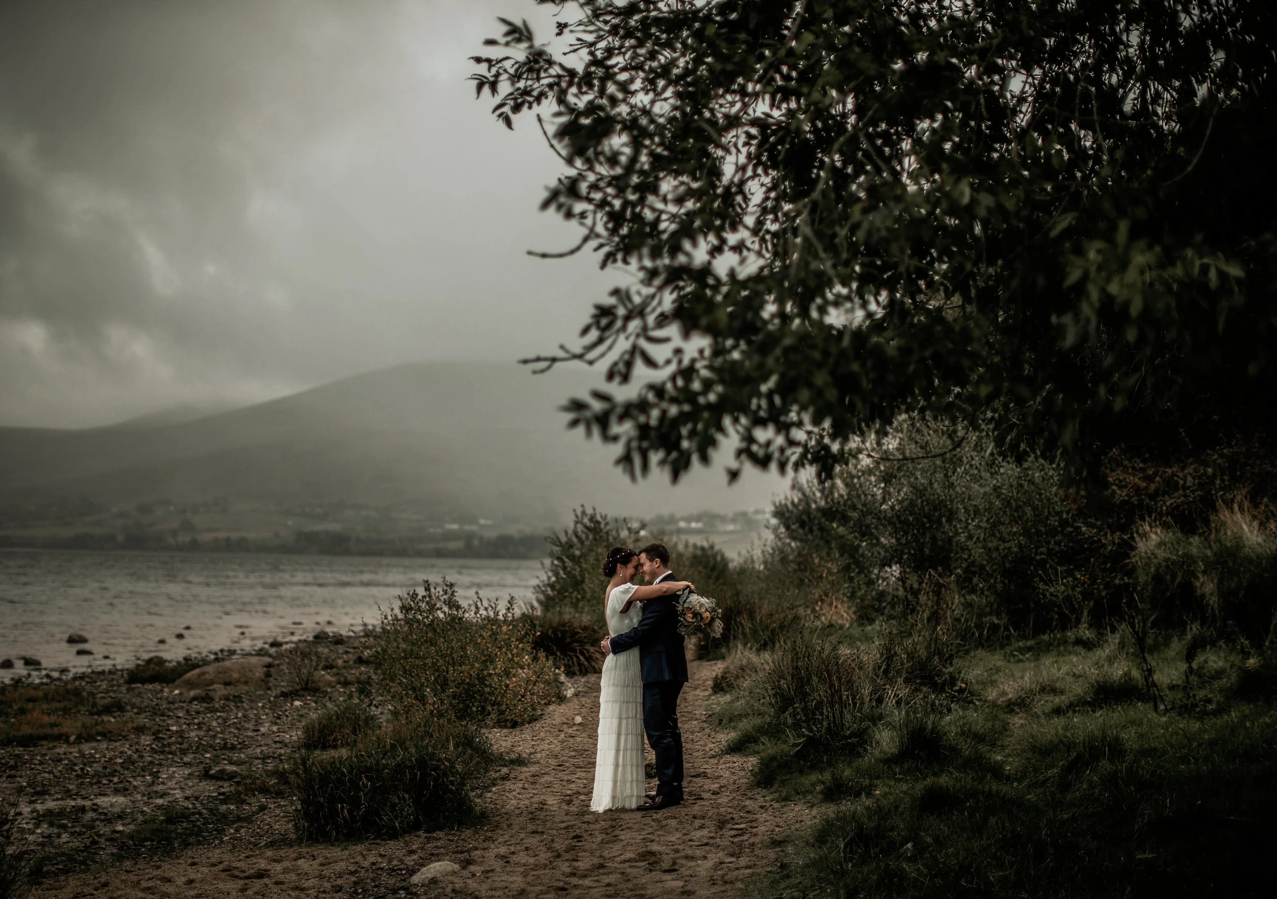 A bride and groom share a romantic embrace on a beachside trail with water and mountains in the background, under a cloudy sky.