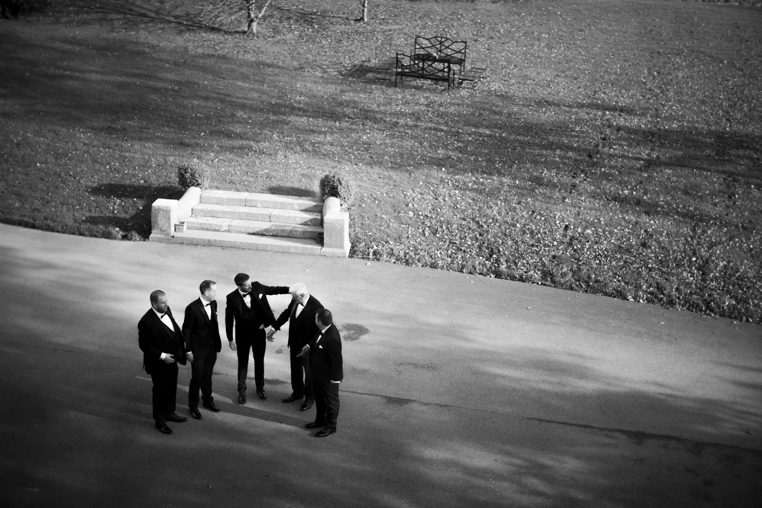 Black and white photo of five men in tuxedos standing on a road in a small circle, talking near a staircase and a grassy area with benches in the background.