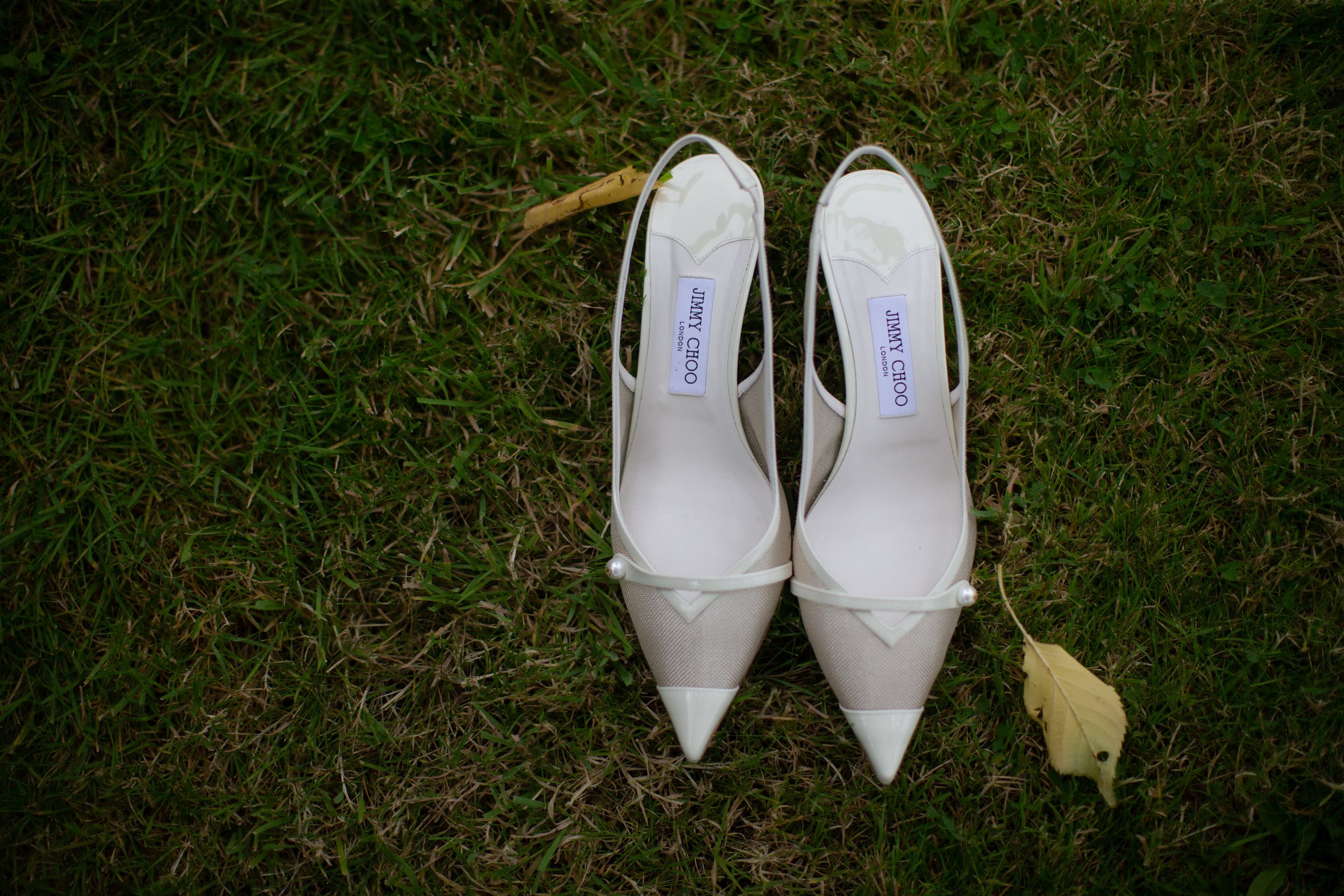 Pair of cream-colored Jimmy Choo high-heeled shoes with pointed toes and pearl embellishments on the straps, placed on green grass with a fallen yellow leaf nearby.