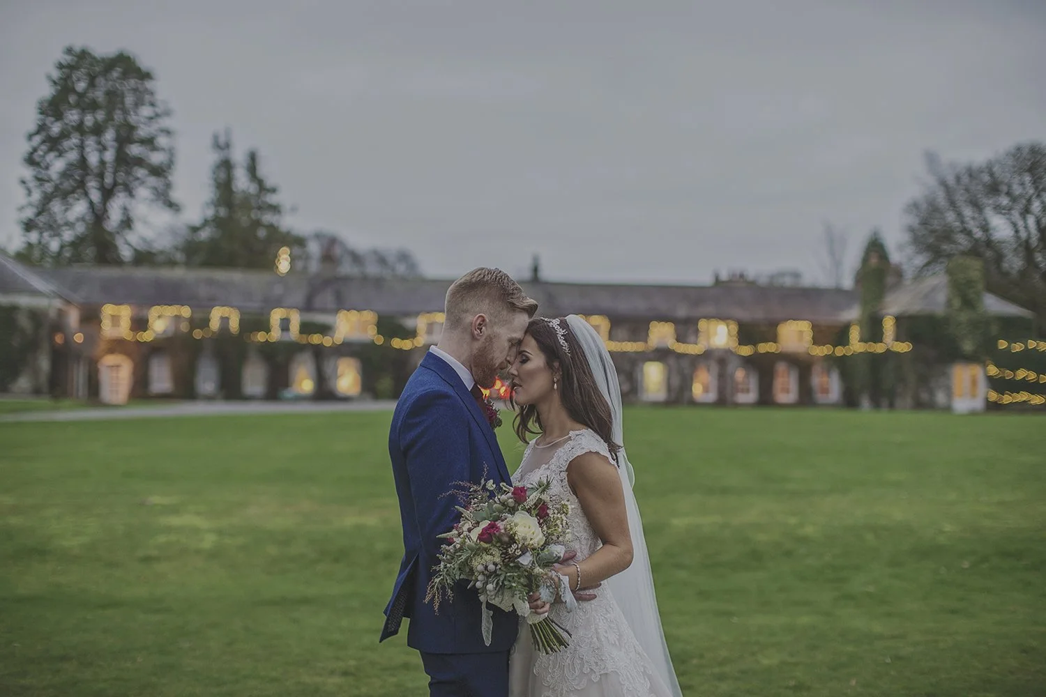 Bride and groom standing close together, forehead to forehead, outdoors on a grassy lawn. The bride holds a bouquet of flowers, and the groom wears a navy blue suit. In the background, there is a large building with string lights, and trees under a c