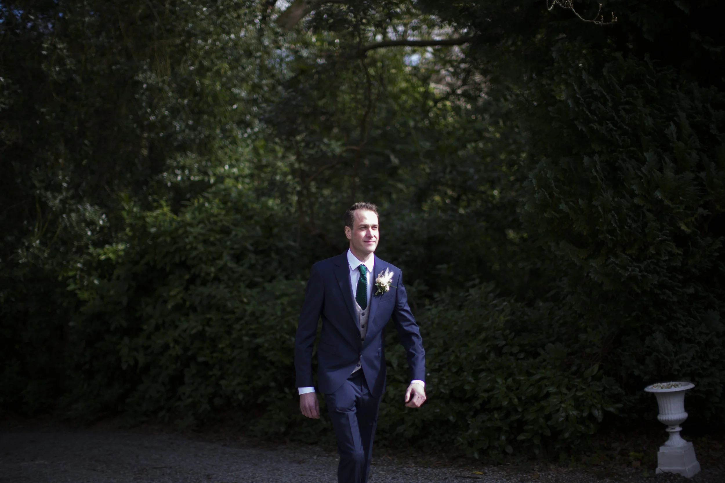 A man in a navy suit with a white shirt and a dark tie walks outdoors in front of green foliage, with a small decorative urn nearby.