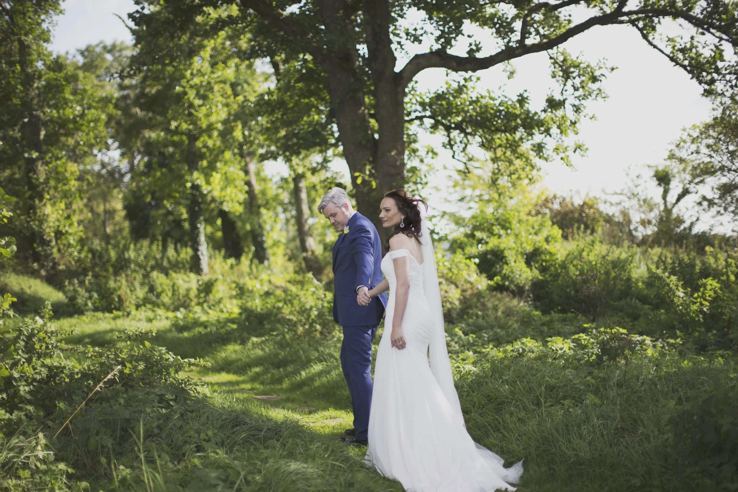 A bride and groom holding hands in a lush green outdoor setting with tall trees and sunlight filtering through the foliage.