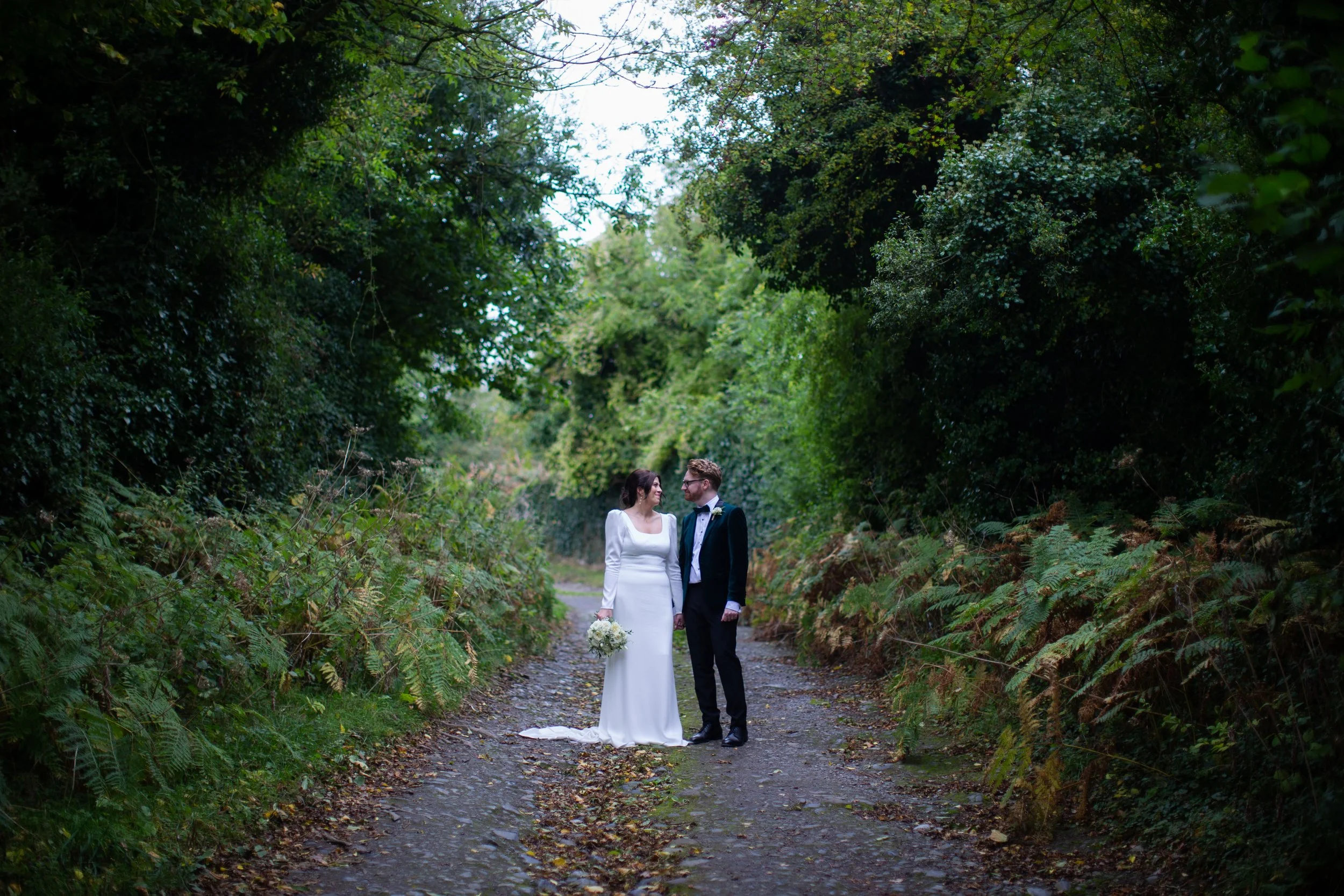 A bride and groom holding hands and looking at each other on a wooded dirt path surrounded by green trees and foliage.