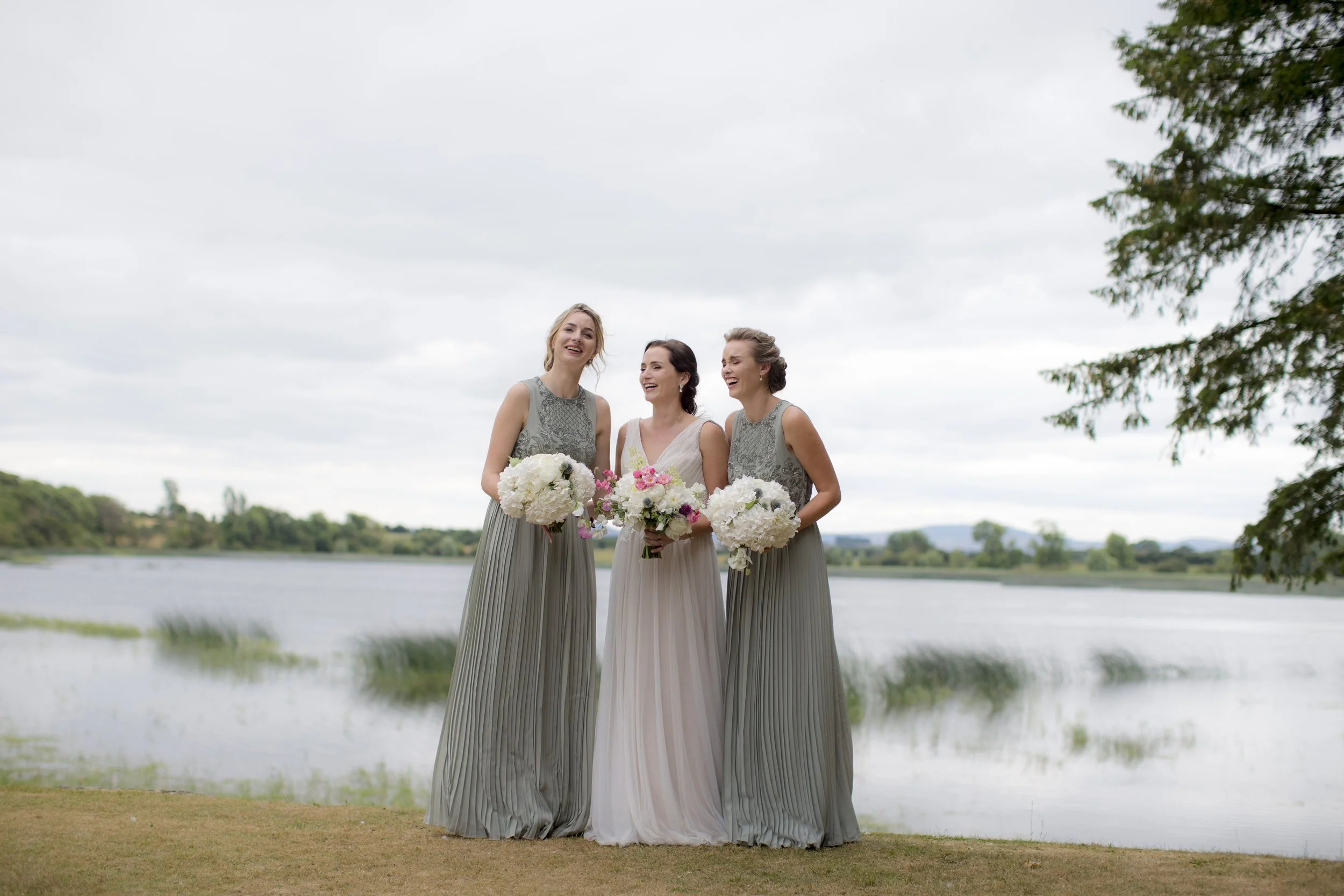 Three women in formal dresses standing outdoors near a lake, holding bouquets of flowers, and smiling.