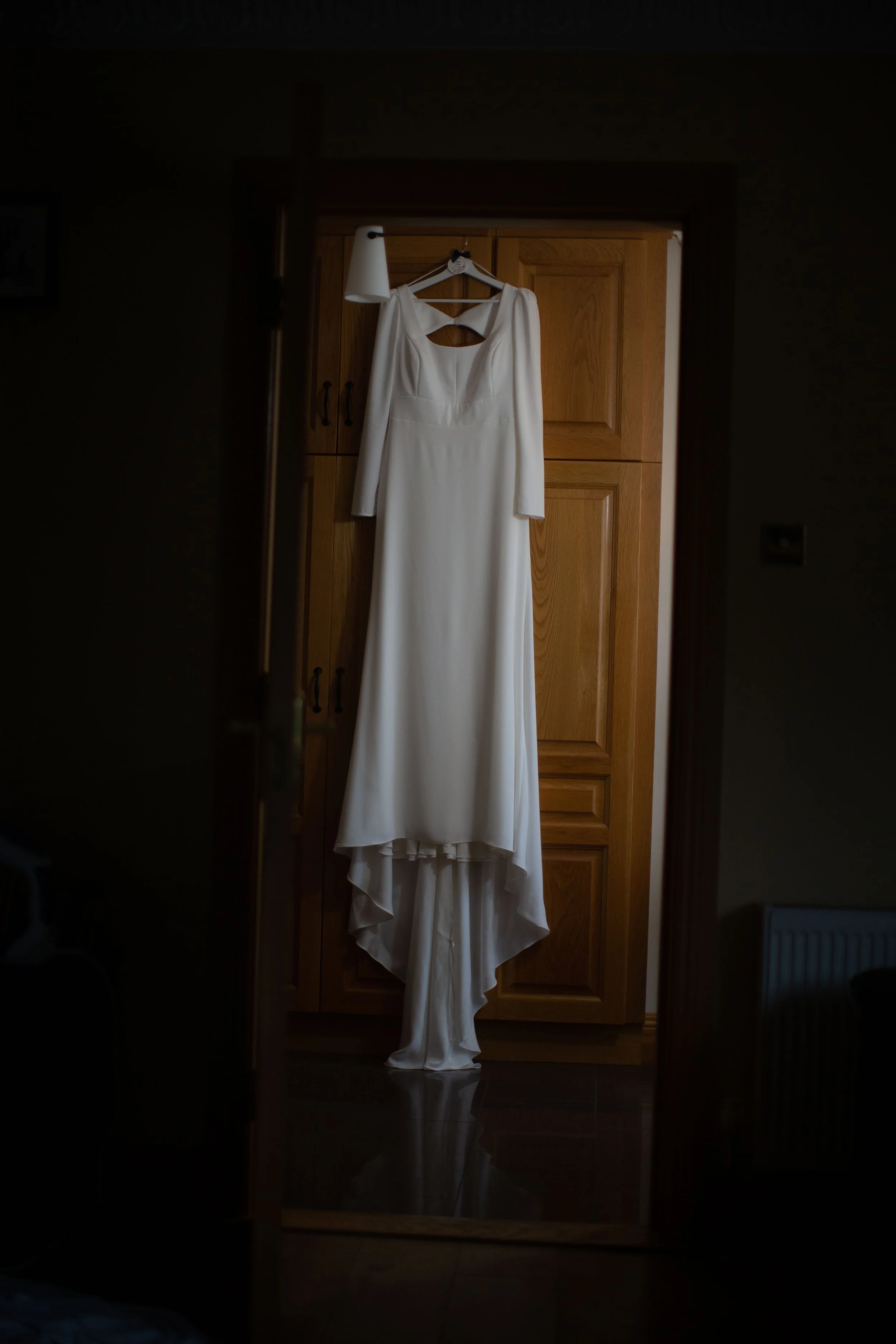 A white wedding dress hanging on a wooden door with a hanger inside a dimly lit room.