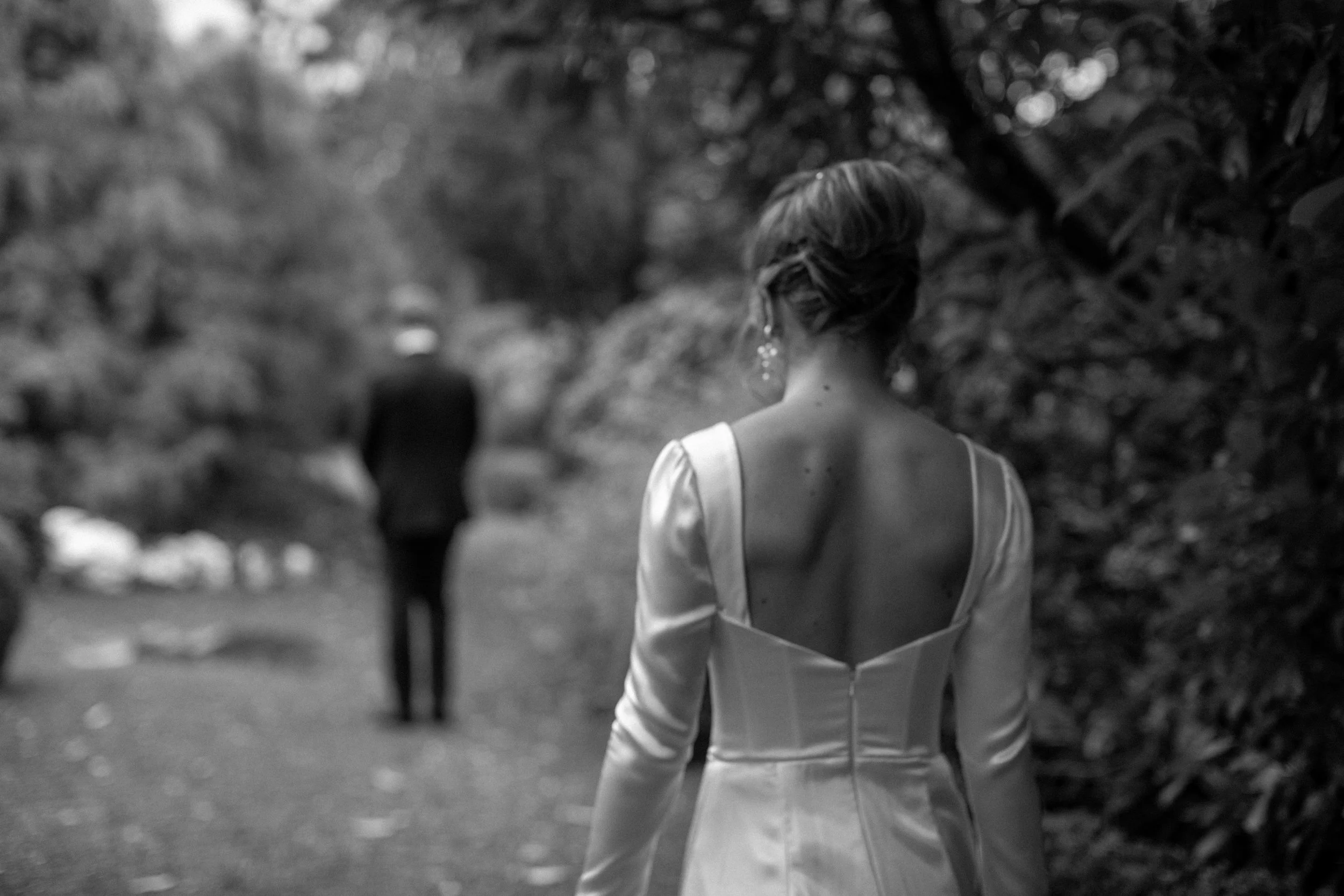 Black and white photo of a woman in a wedding dress with back to the camera, walking on a forest trail away from a man in a suit standing in the background.