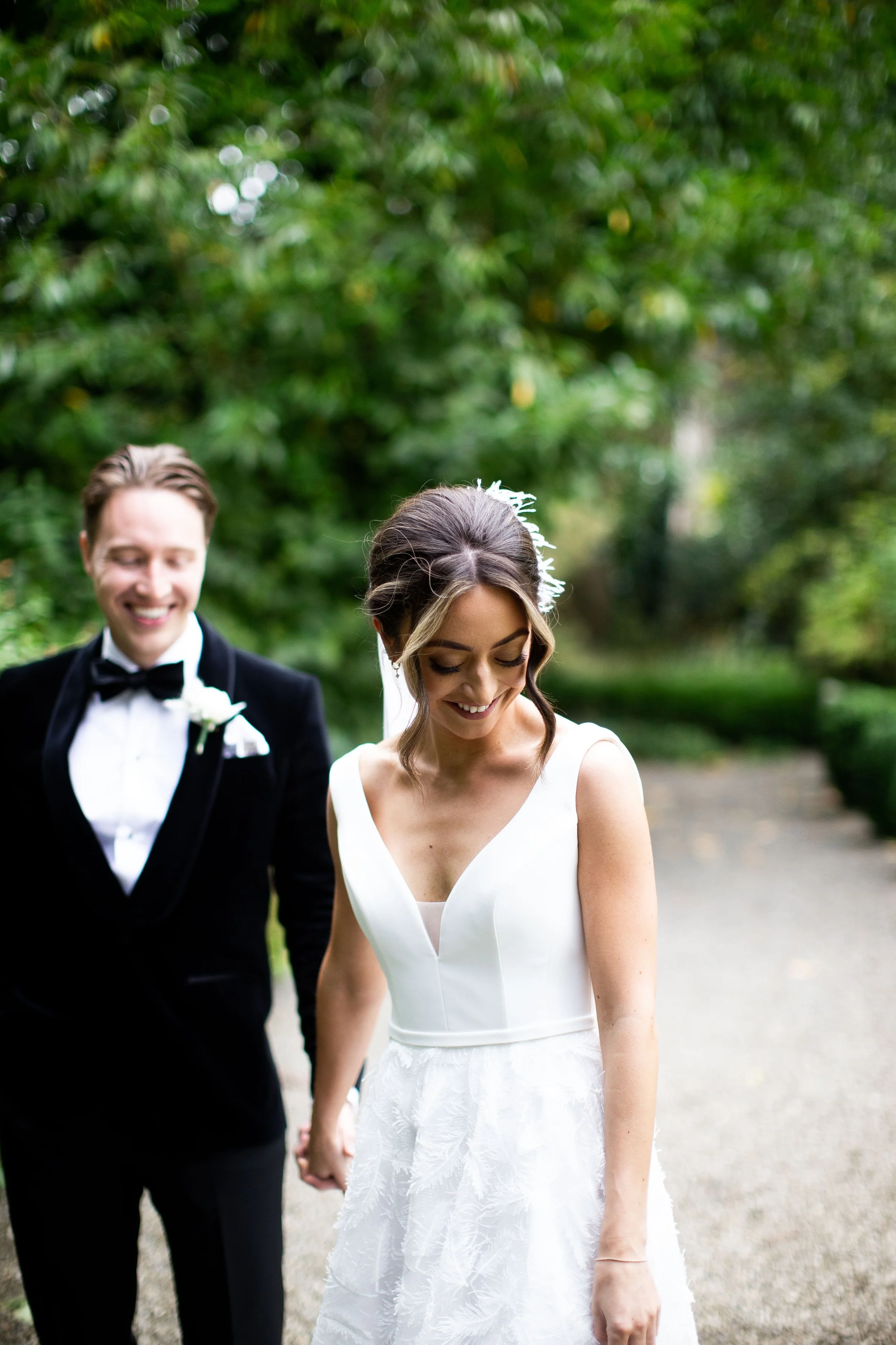 A bride and groom holding hands, walking outdoors on a pathway surrounded by greenery, smiling, and dressed in wedding attire.