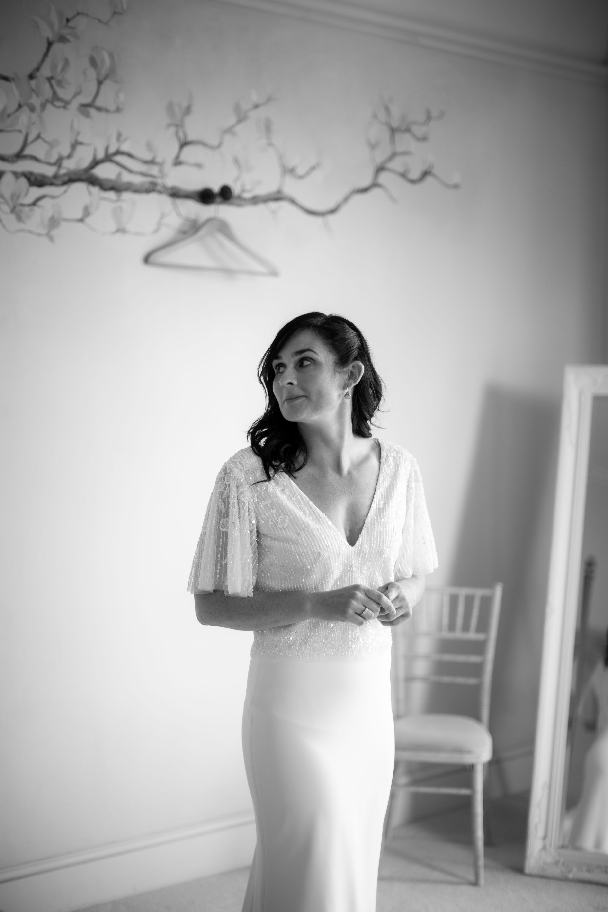 A woman in a wedding dress standing indoors with a mirror and a chair in the background, black and white photograph.