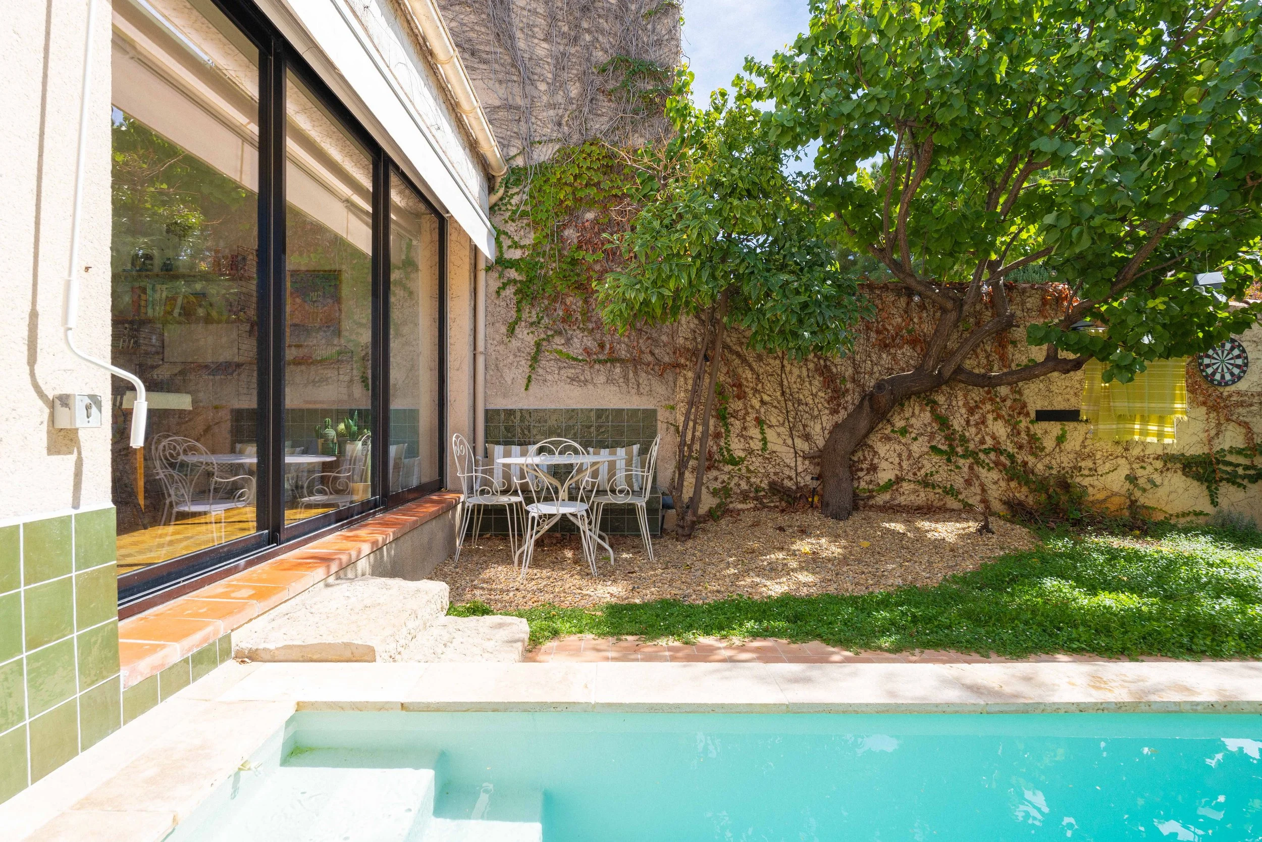 Une cour intérieure avec un arbre, une table en métal blanc et des chaises, une piscine en premier plan, et une façade de maison avec de grandes fenêtres.