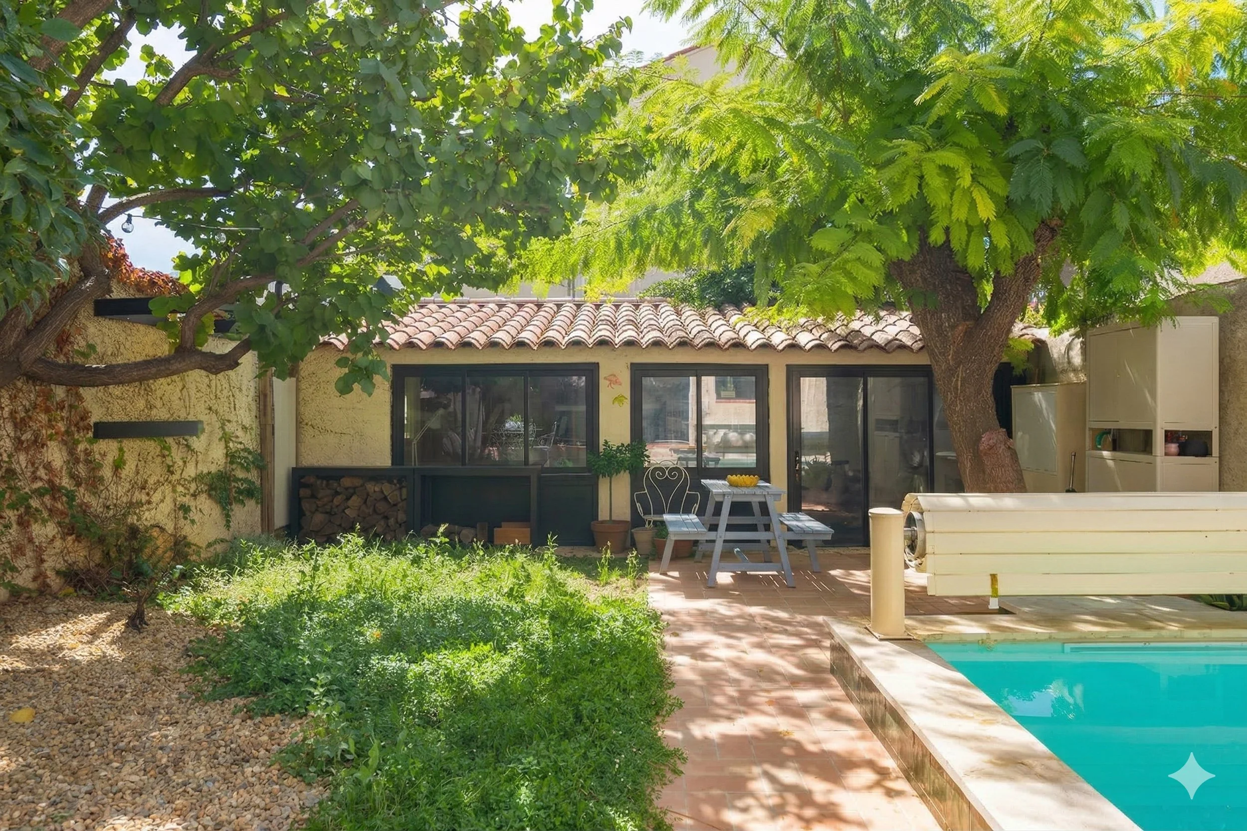 Coin de jardin avec terrasse, piscine, arbre ombrageant la zone, mobilier d'extérieur, bâtiment avec fenêtres et porte en arrière-plan.