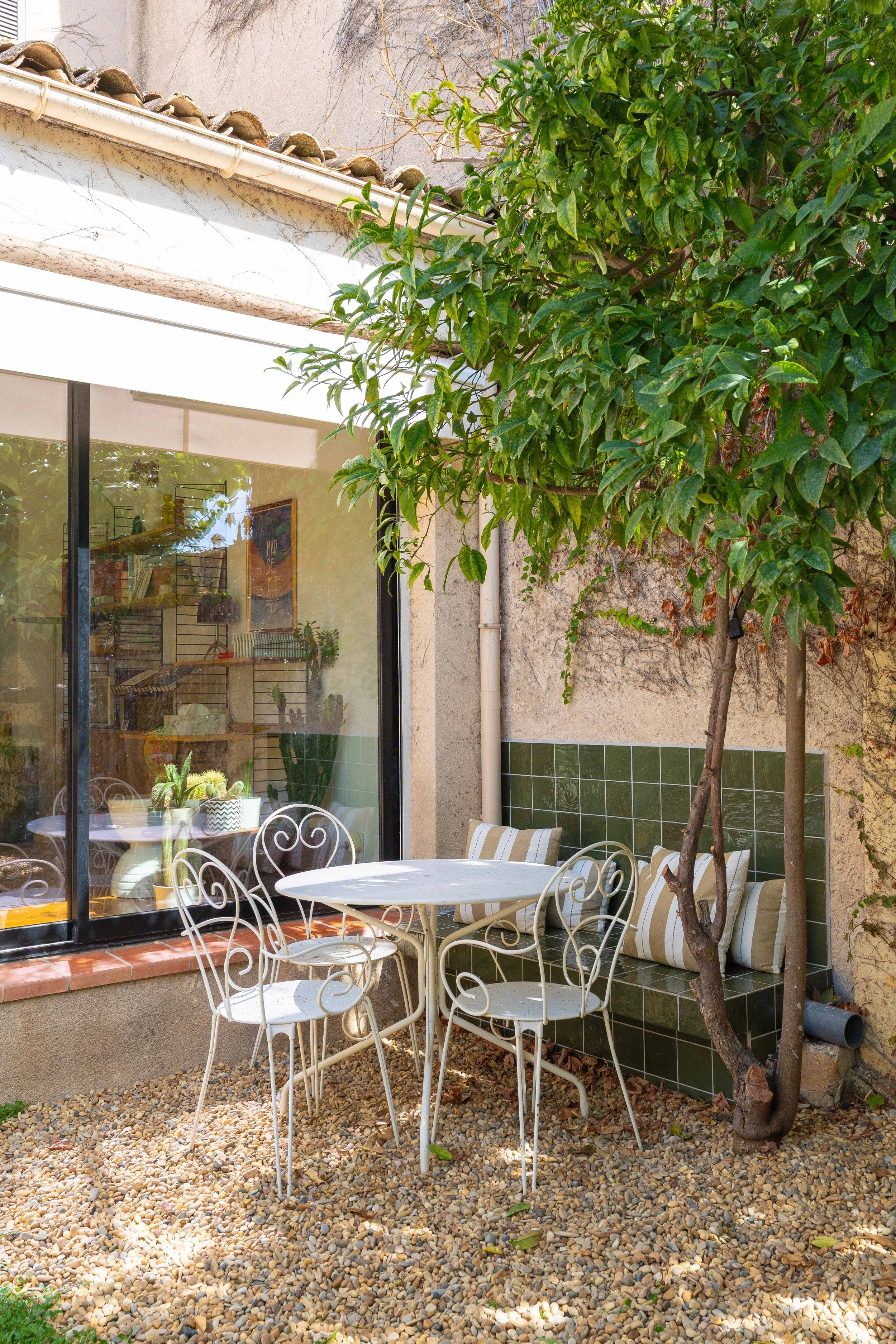 Un coin patio avec une table ronde en métal blanc, entourée de cinq chaises en métal blanc au sol composé de petits galets. Un arbre avec des feuillages verts offre de l'ombre. À l'arrière, une banquette en céramique verte avec des coussins rayés cou
