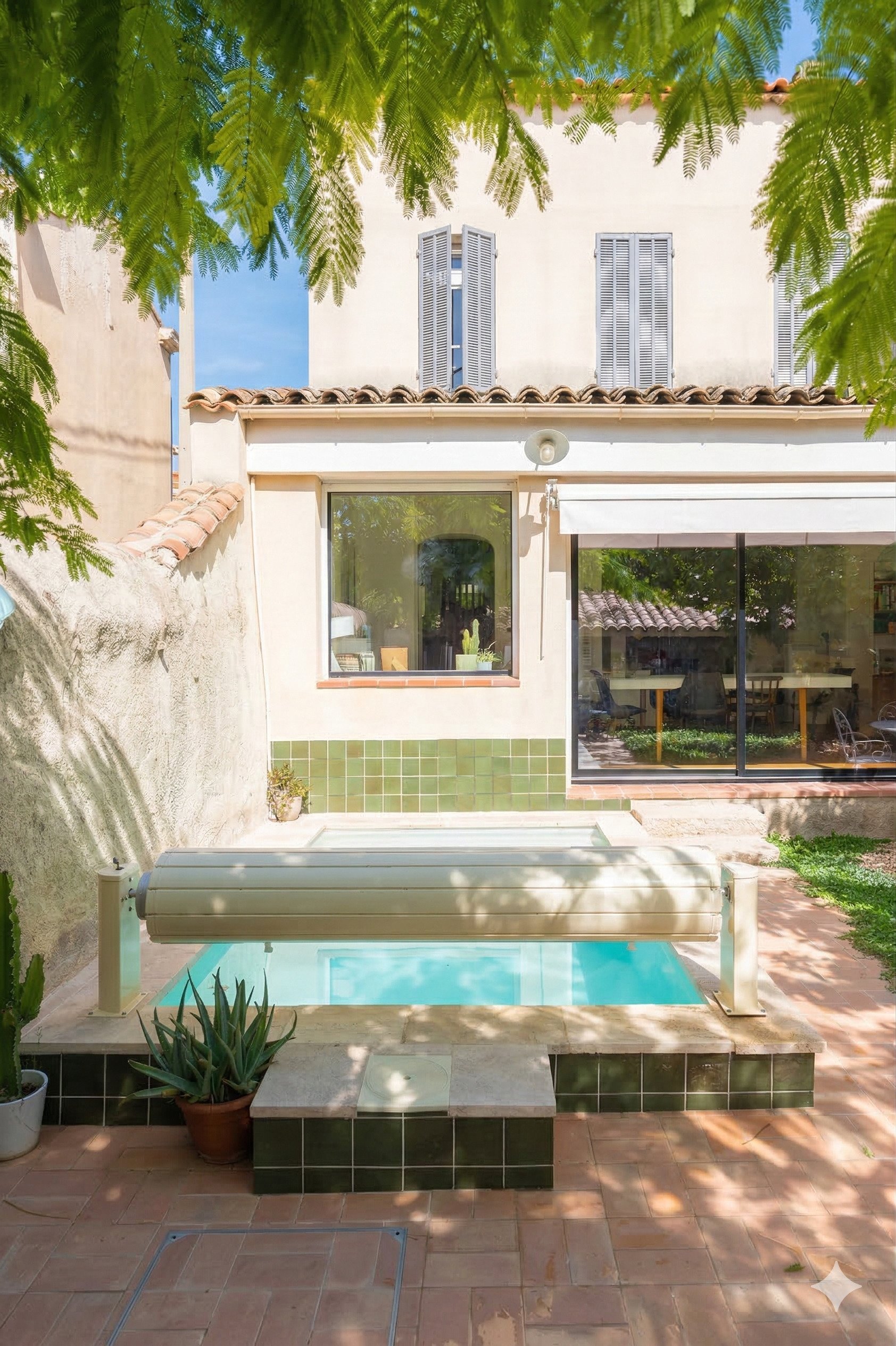 Cour arrière avec piscine, plantes en pot, murs blancs, fenêtres avec volets gris, terrasse en tuiles, ombres d'arbres, maison avec toit en tuiles, ambiance ensoleillée.