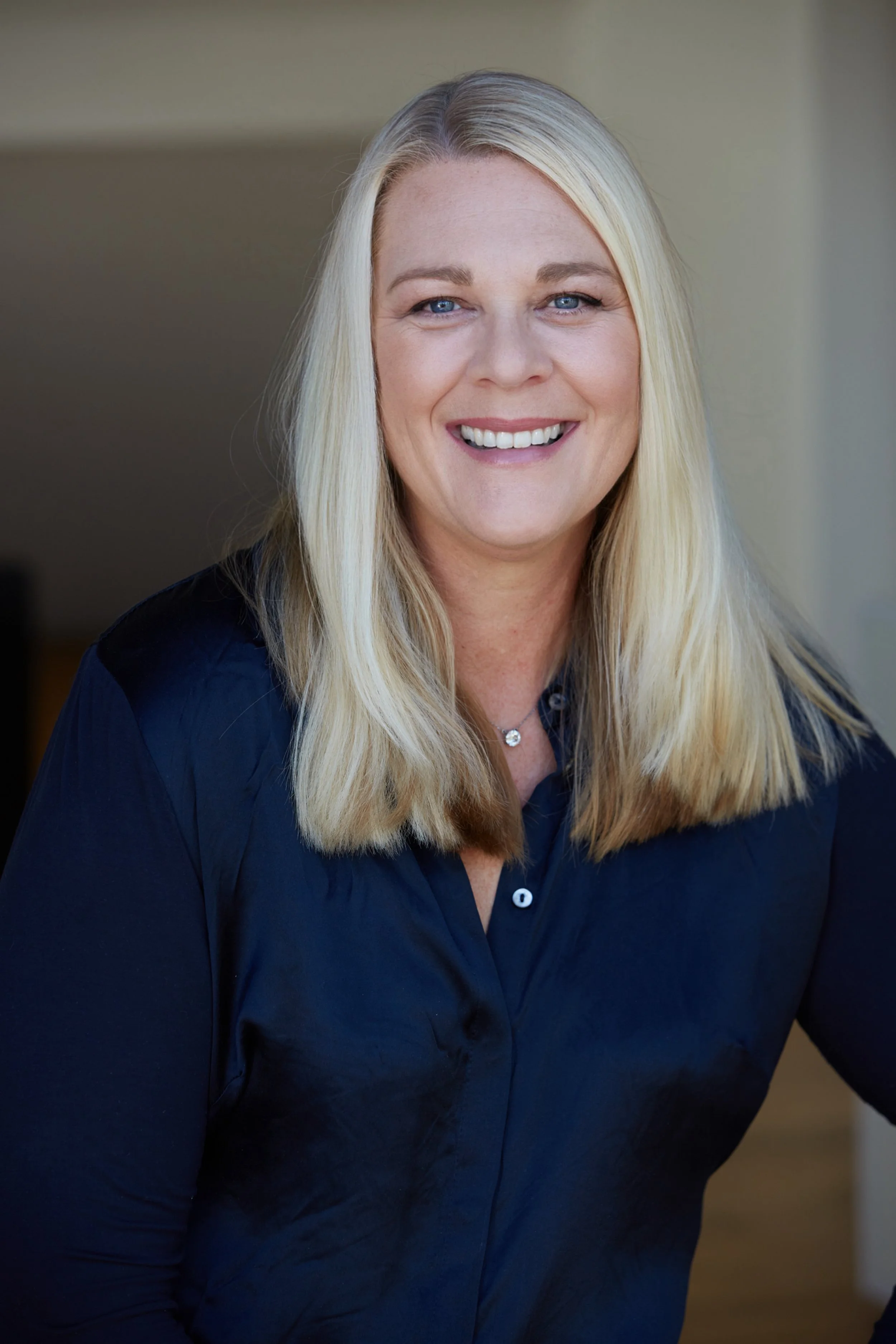 A smiling woman with long blonde hair wearing a navy blue shirt, indoors.