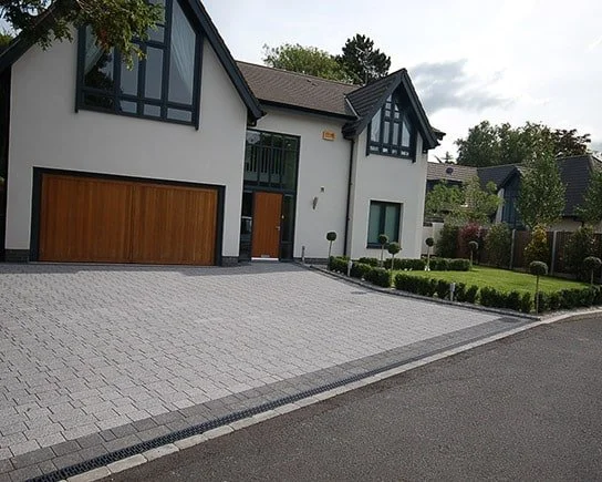 Modern two-story house with a gray driveway, wooden garage door, and large windows, with a green lawn and trees in the background.