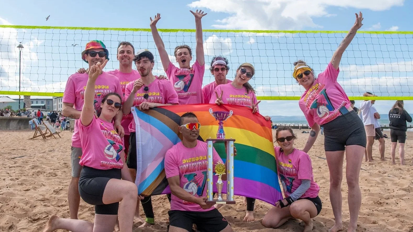 A volleyball team pose as a group on porty beach behind a pride flag and a large trophy