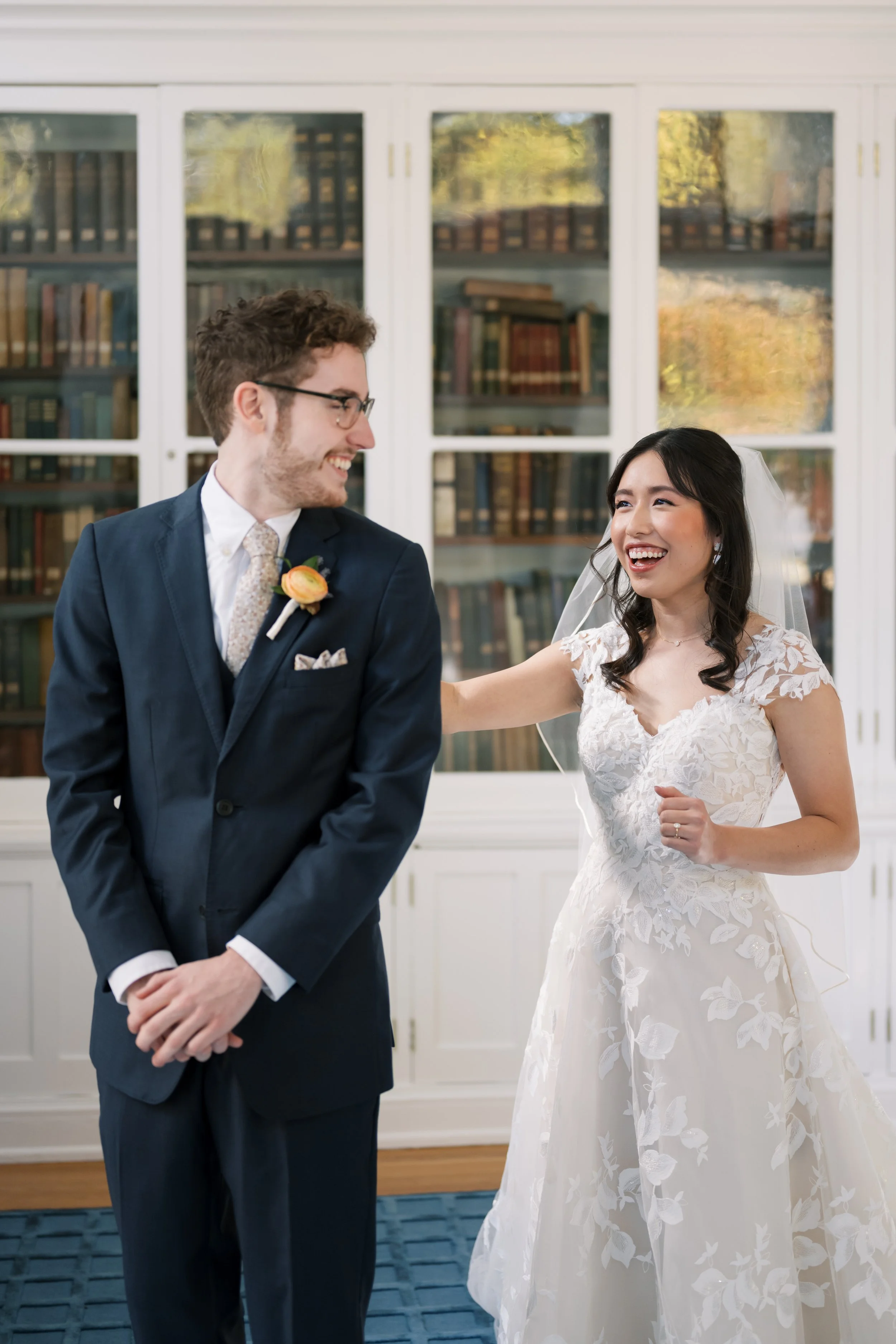 Woman in white, floral wedding dress and veil tapping man in darky navy suit on shoulder during a wedding first look in front of glass encased bookshelves