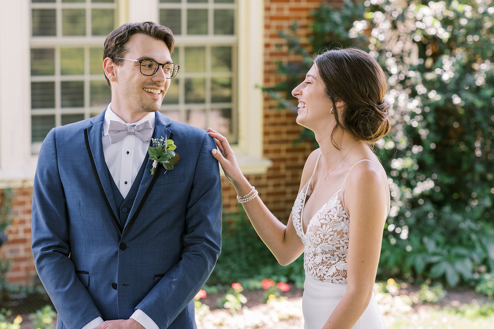 Man in navy suit on left looking at woman on right in spaghetti strap wedding dress smiling with her hand on man's shoulder. Both are standing in front of a brick building.