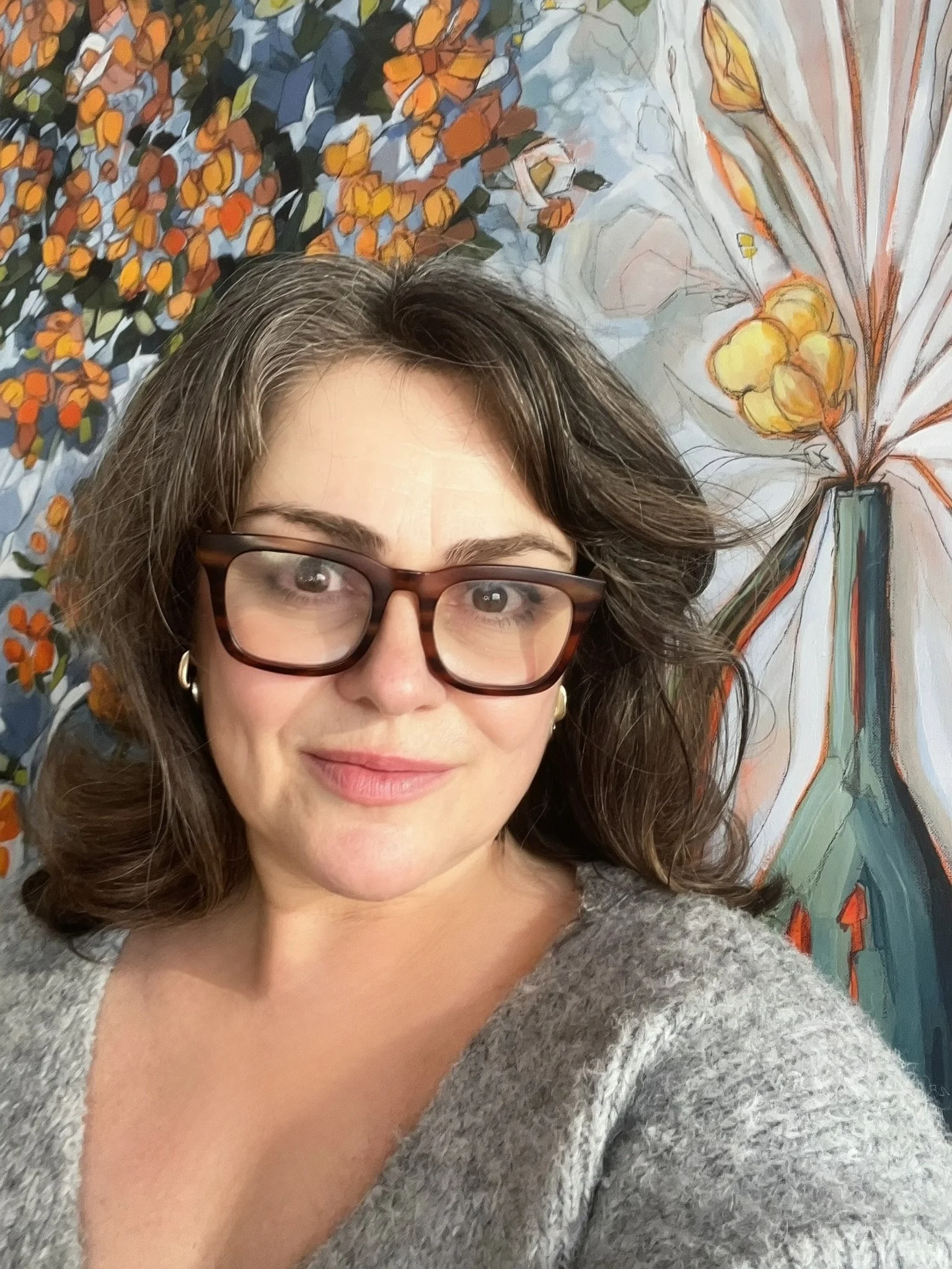 A woman, Christie Baker, wearing glasses and a grey sweater, smiling with dark wavy hair, in front of her still life painting, abstract-patterned background.