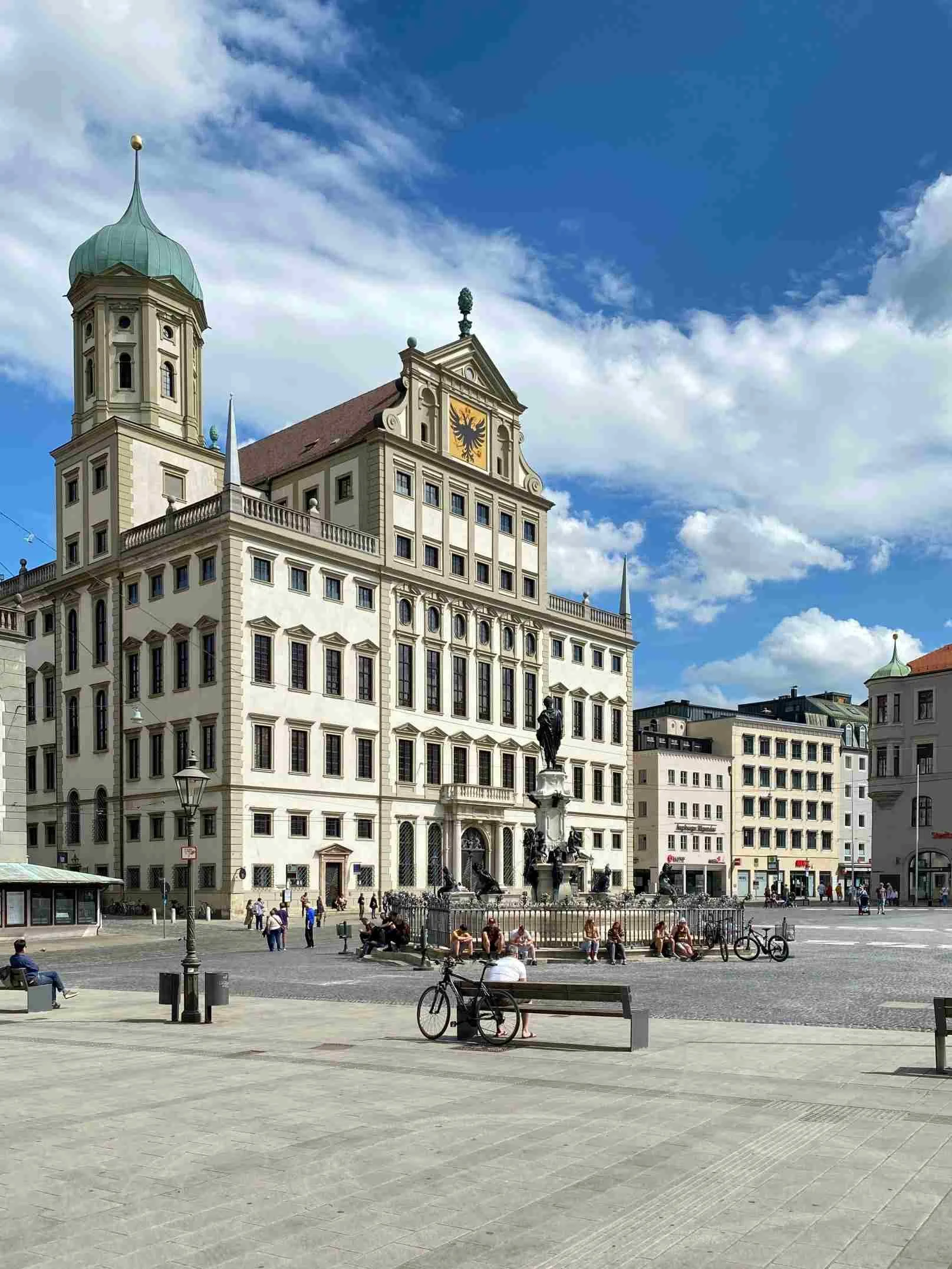 Blick auf ein historisches Gebäude mit einer Uhr im Zentrum eines belebten Platzes. Es gibt Menschen, Bänke, Fahrräder und eine Statue im Vordergrund. Der Himmel ist blau mit Wolken.