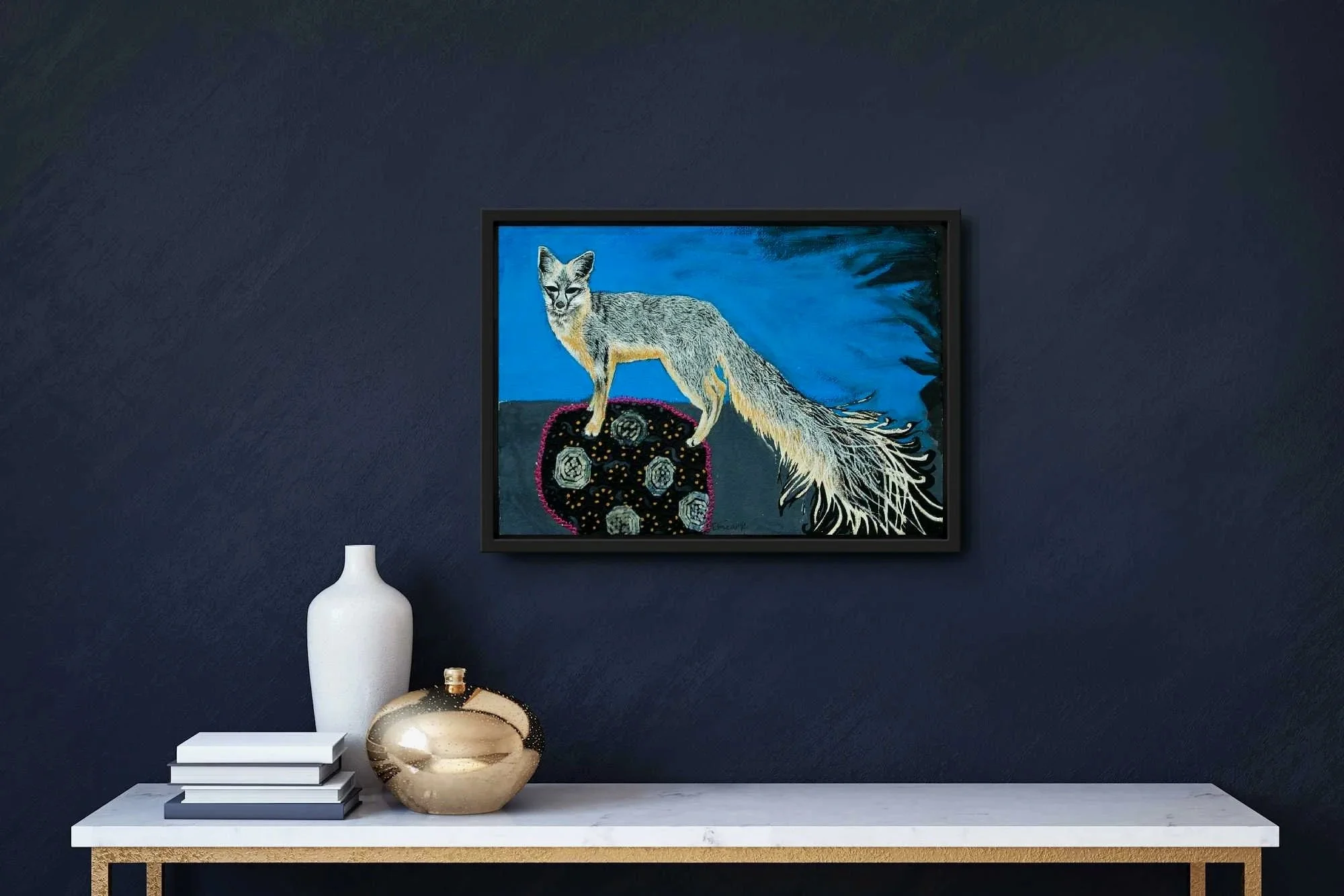 Framed painting of a fox with a long bushy tail standing on a rock against a blue sky, on a navy blue wall. Below the painting, a white vase, a round gold decorative object, and a stack of four books are placed on a white marble table.