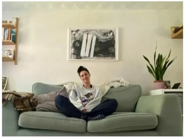 Person sitting cross-legged on a light-colored sofa in a room with minimal decor, including a framed abstract picture, potted plant, and shelves with books.